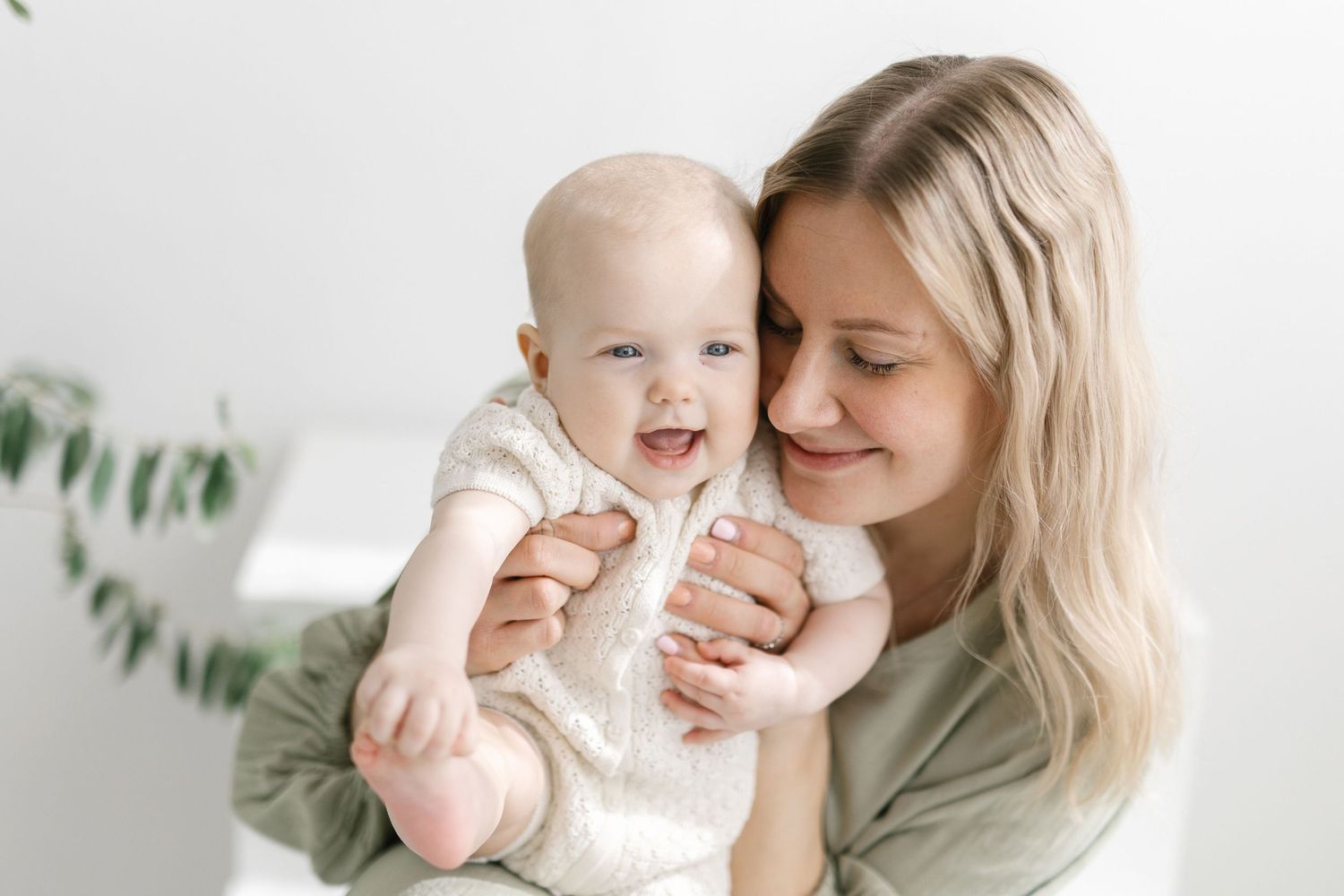A smiling mother with blonde hair holds her happy laughing baby in a bright white room.