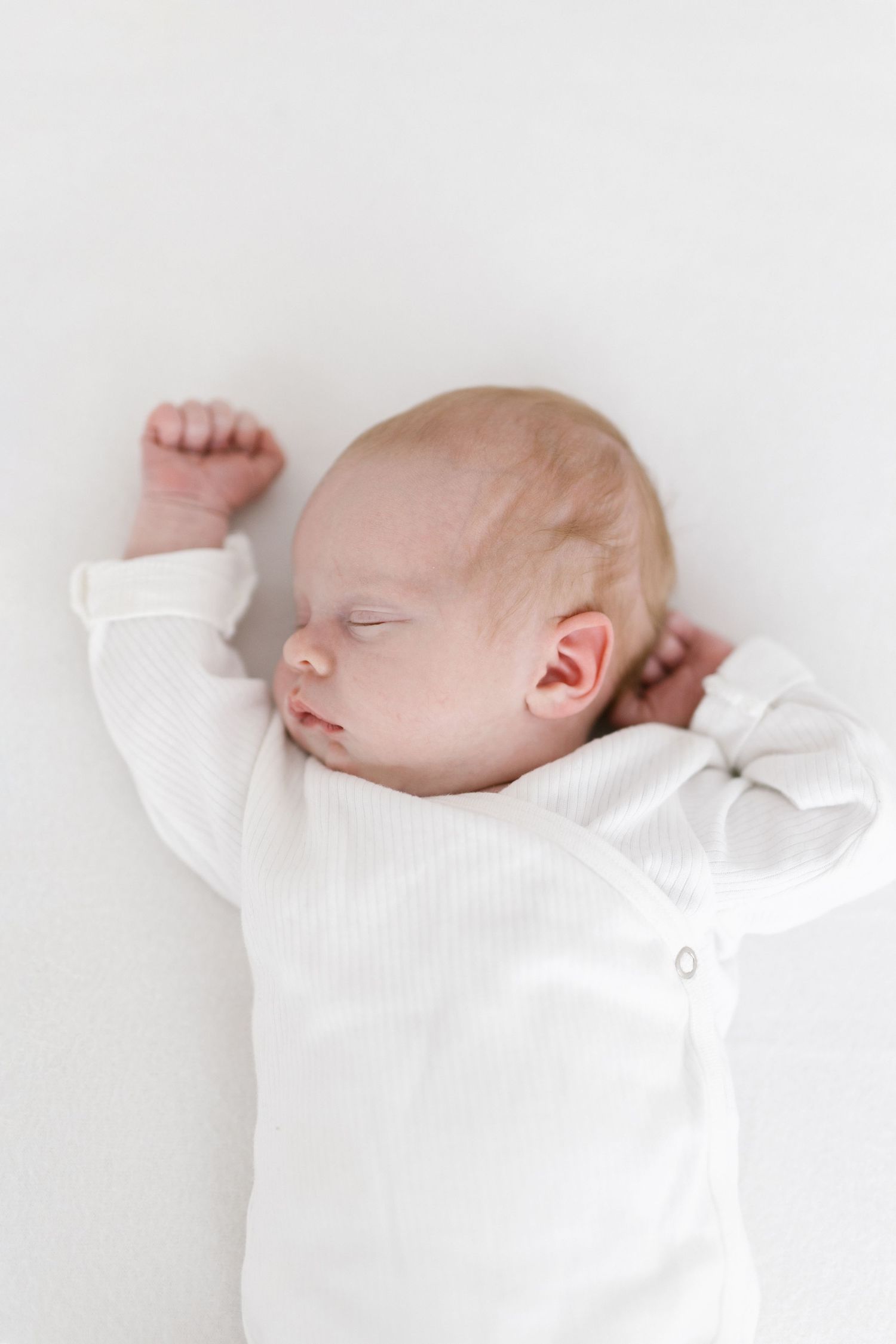 Peaceful sleeping newborn in white onesie against light background.