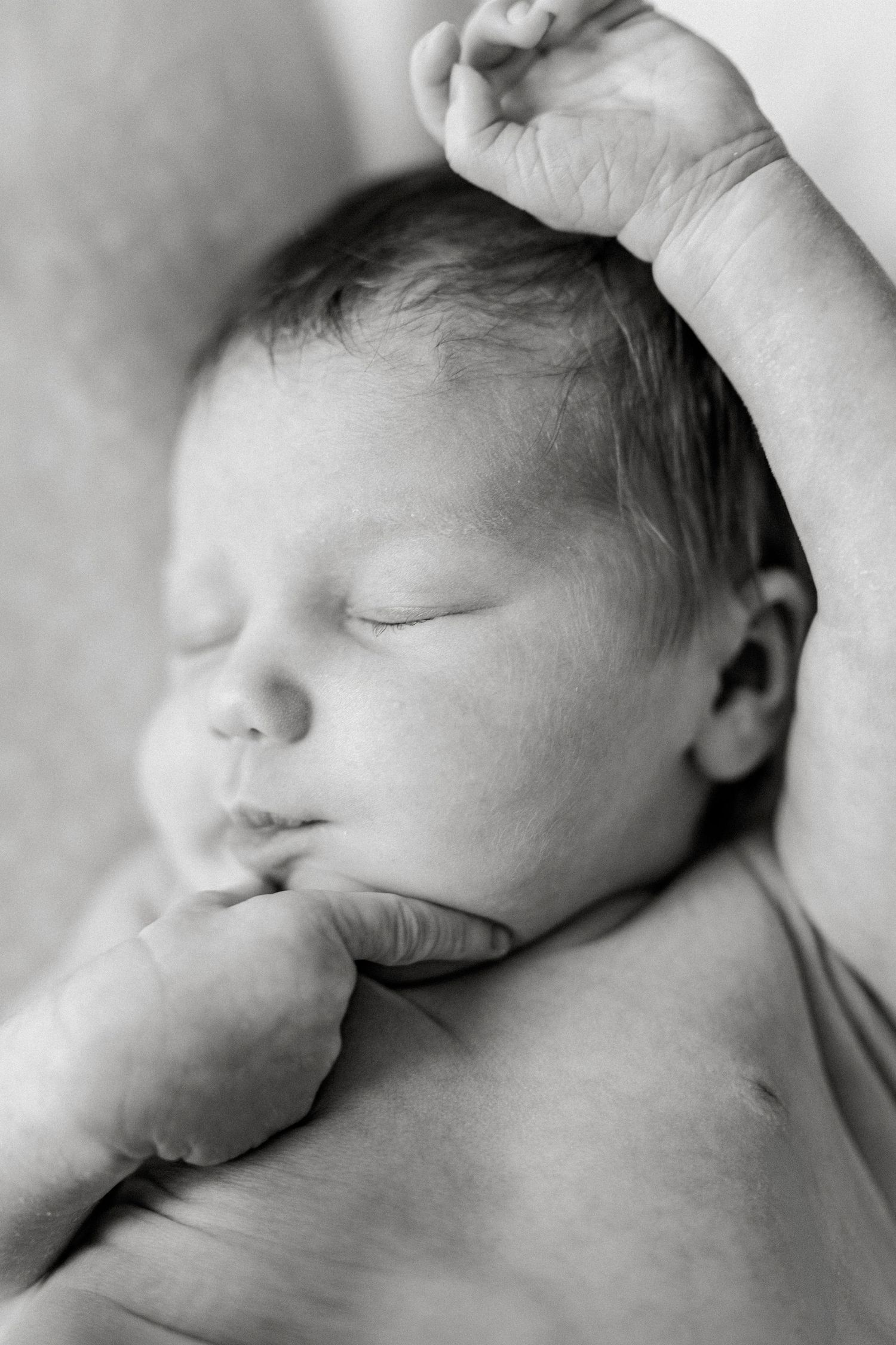 Black and white photo of a sleeping newborn baby with tiny fists raised, peaceful expression on soft blanket.