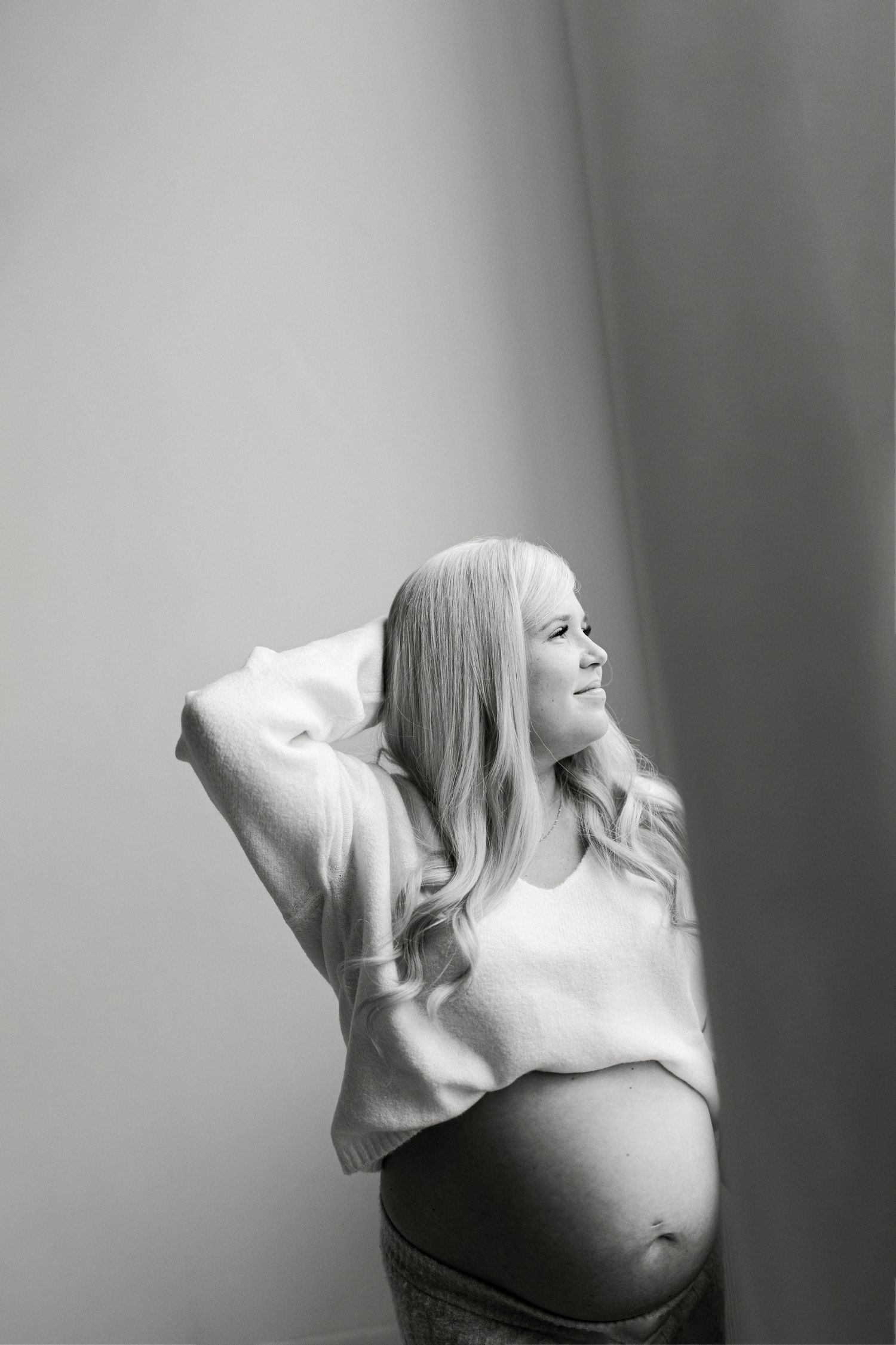 Black and white maternity photo of a blonde pregnant woman in a white sweater, smiling and posing gracefully.