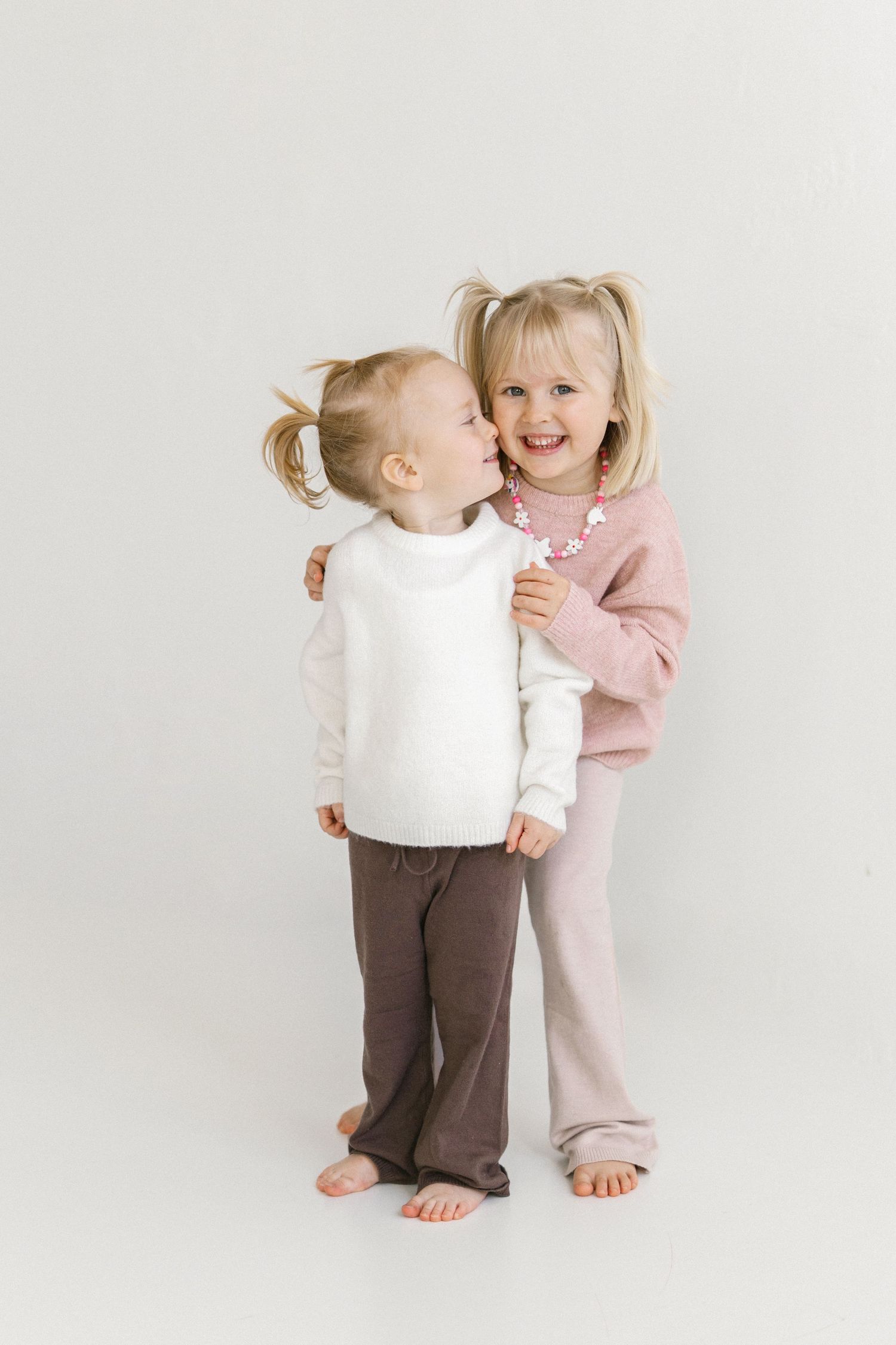 Two young blonde girls in pastel outfits share a sweet kiss moment against a white background.