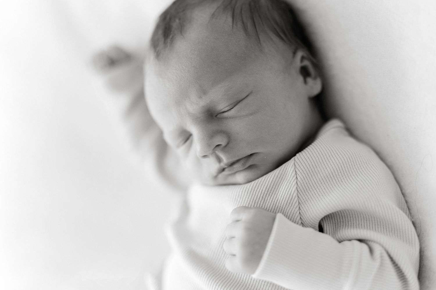 Black and white newborn baby sleeping peacefully, wearing a white onesie, with tiny fist raised near face.
