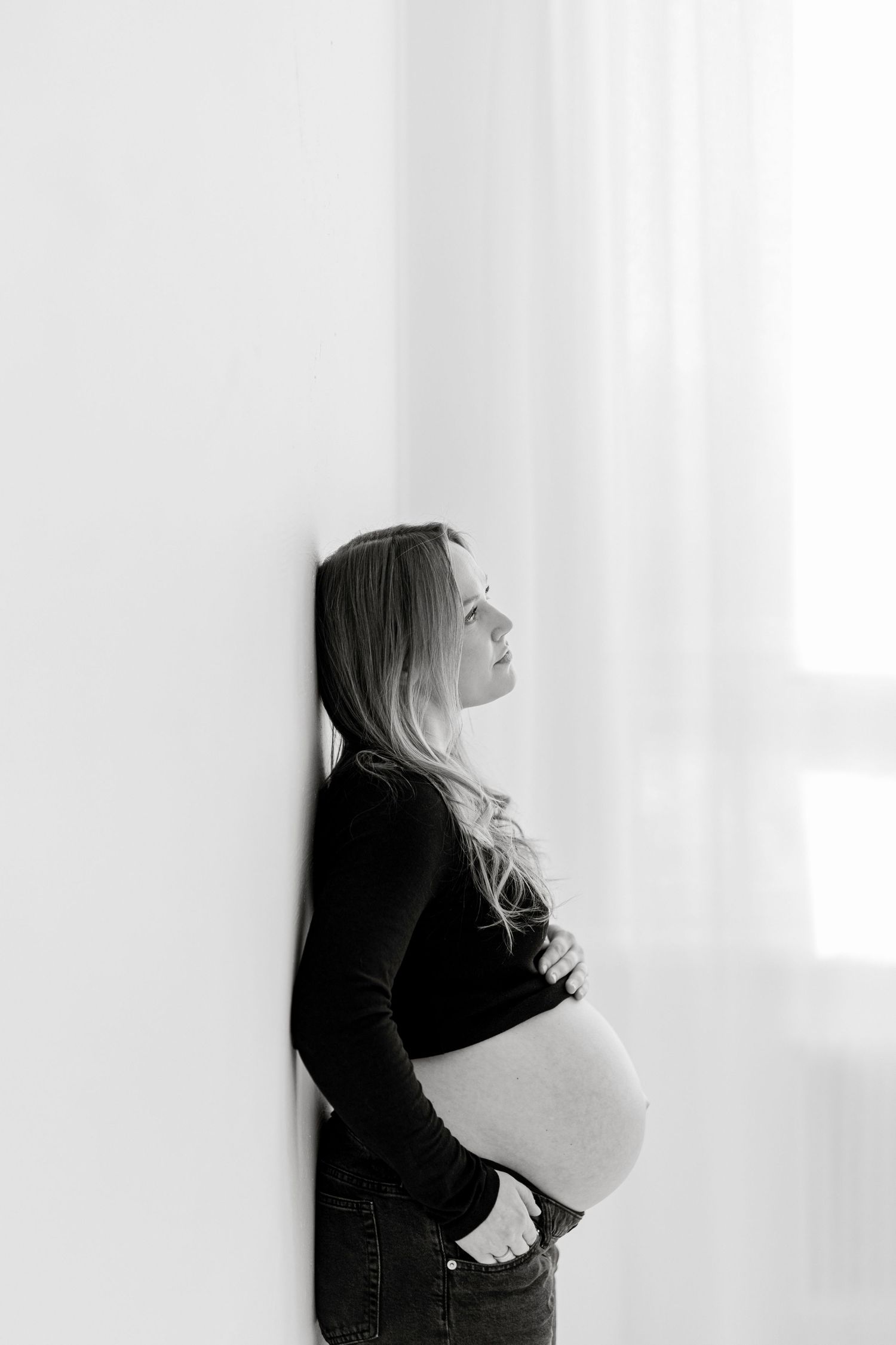 Black and white photo of a pregnant woman leaning against a wall, cradling her baby bump while gazing upward.
