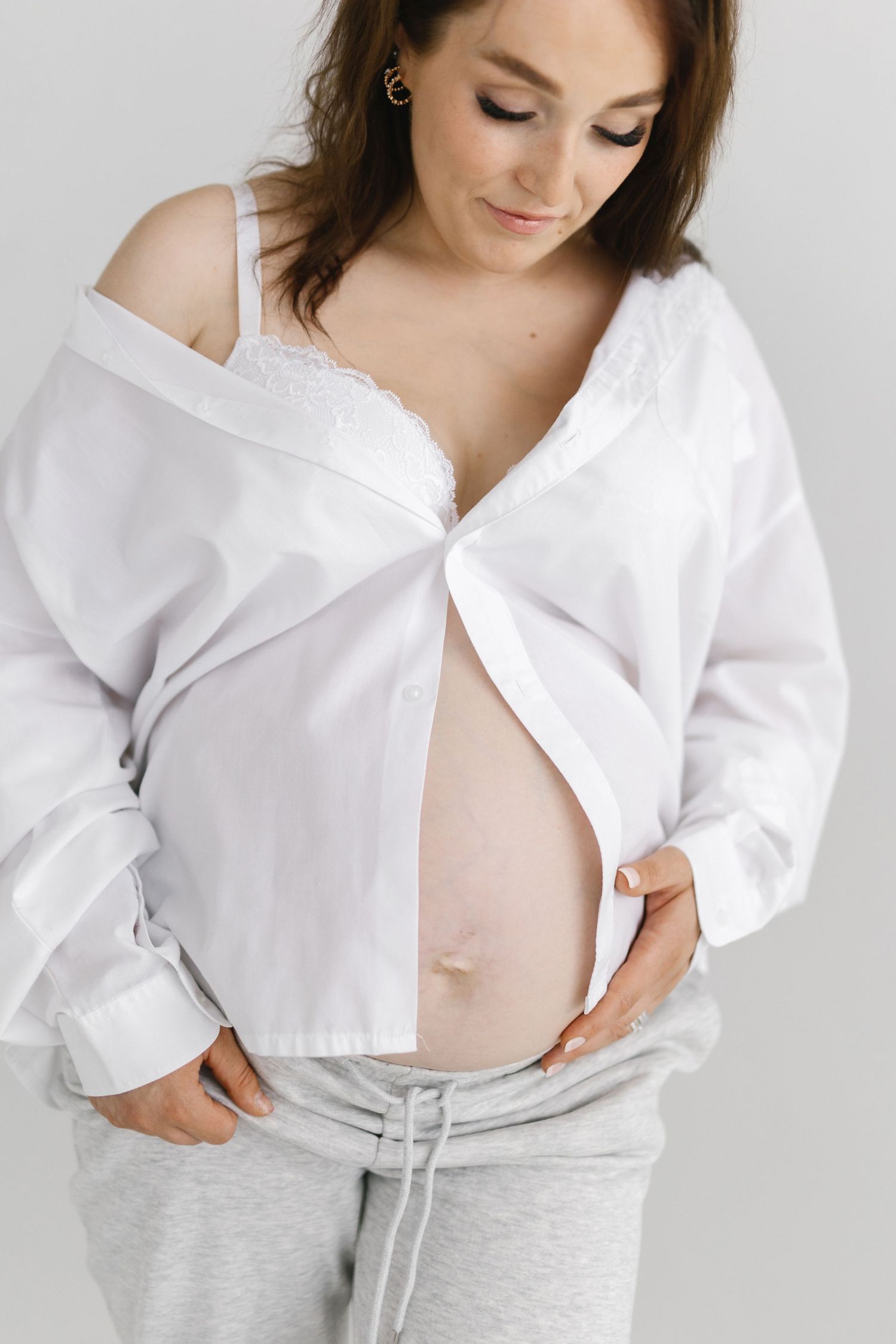 Woman in white button-down shirt and gray sweatpants shows pregnant belly.