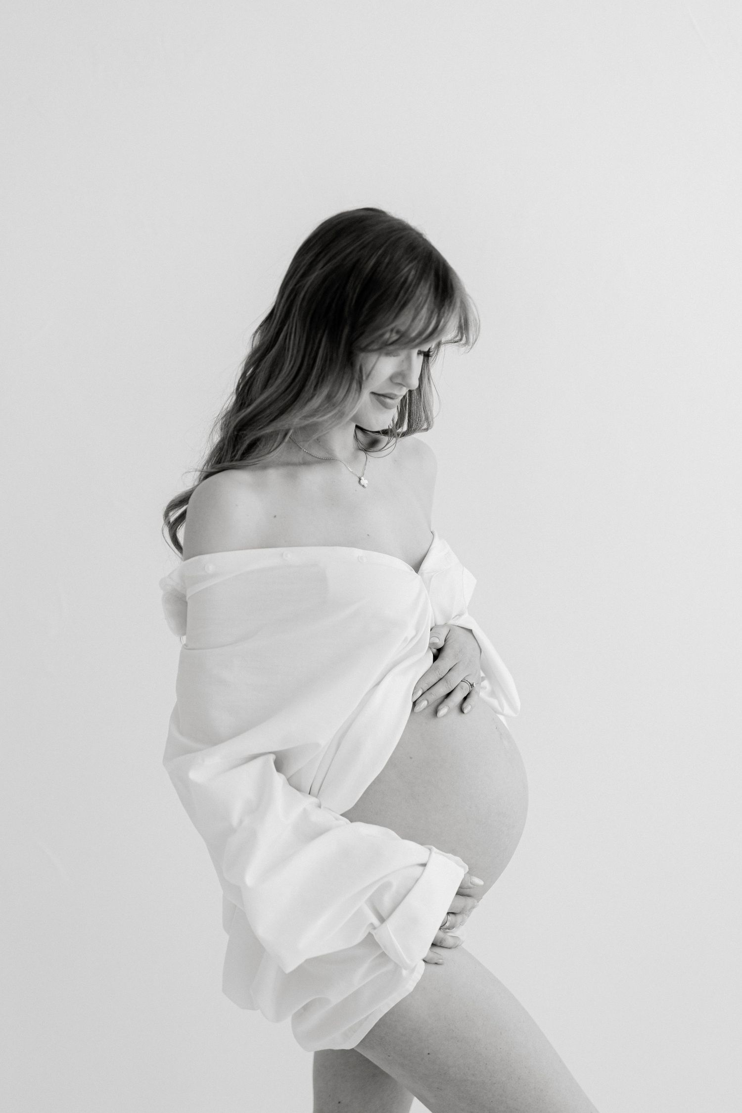 Black and white maternity photo of a pregnant woman in an off-shoulder white shirt cradling her baby bump.