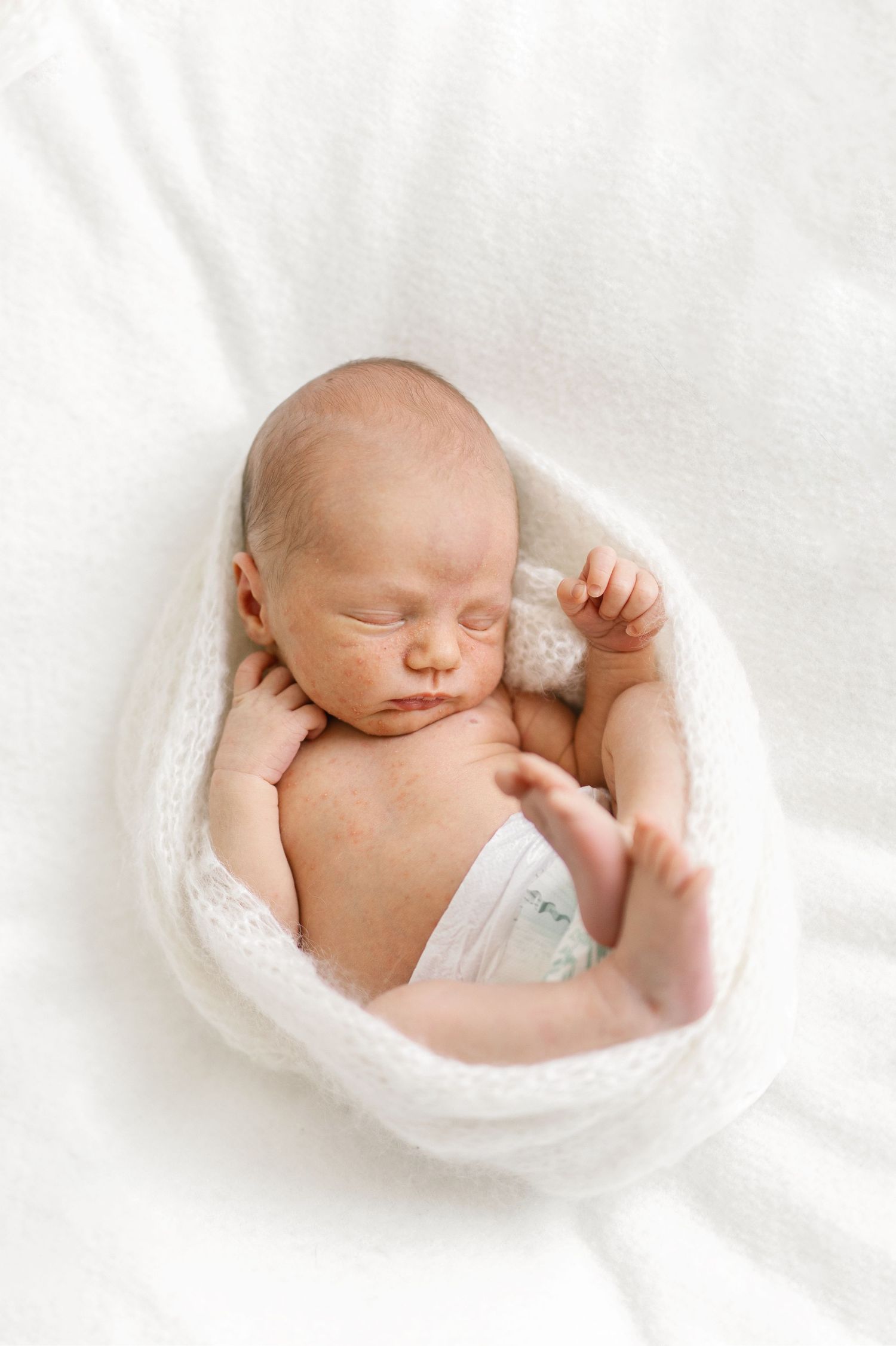 Peaceful newborn baby sleeping wrapped in a white knit blanket on a soft white background.