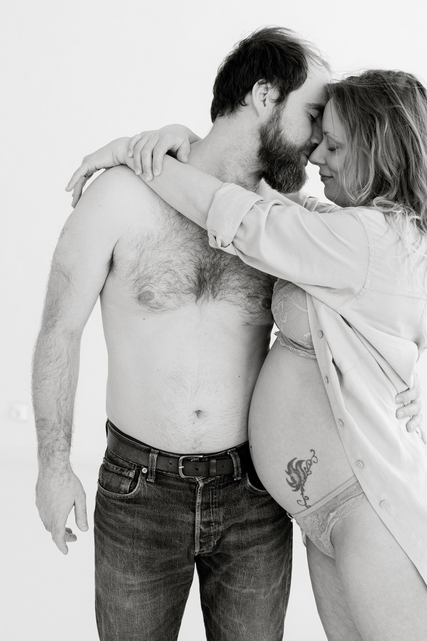 Black and white maternity photo of shirtless man and pregnant woman in lingerie embracing tenderly.