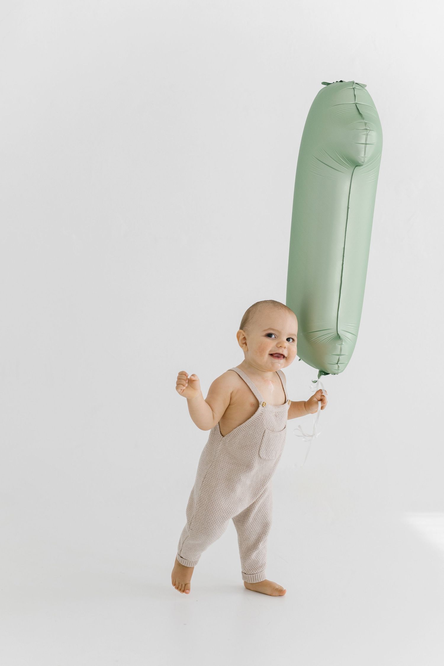 Baby in beige romper plays with mint green balloon on white background.
