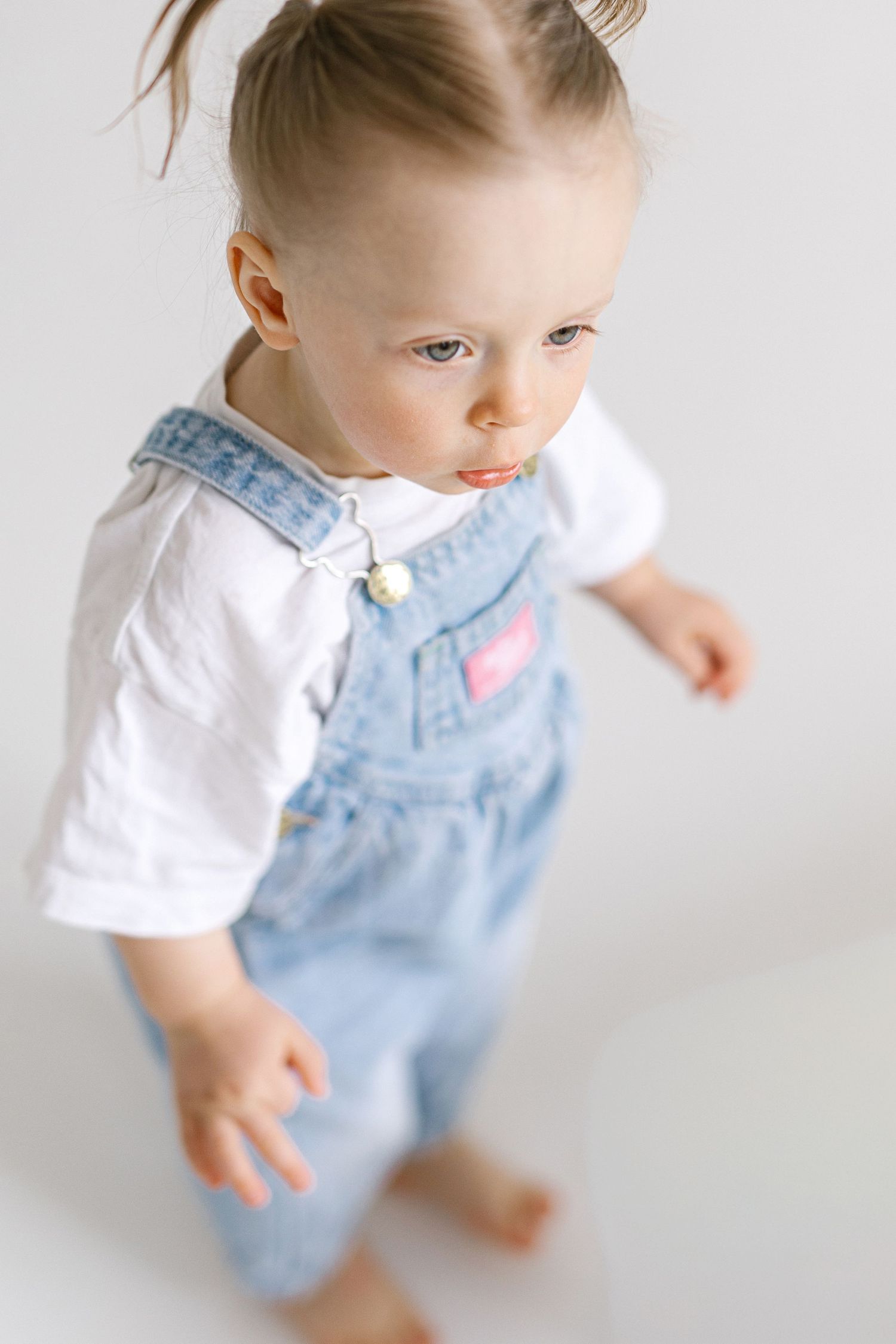 Toddler girl in denim overalls and white shirt looks down while standing barefoot against white background.