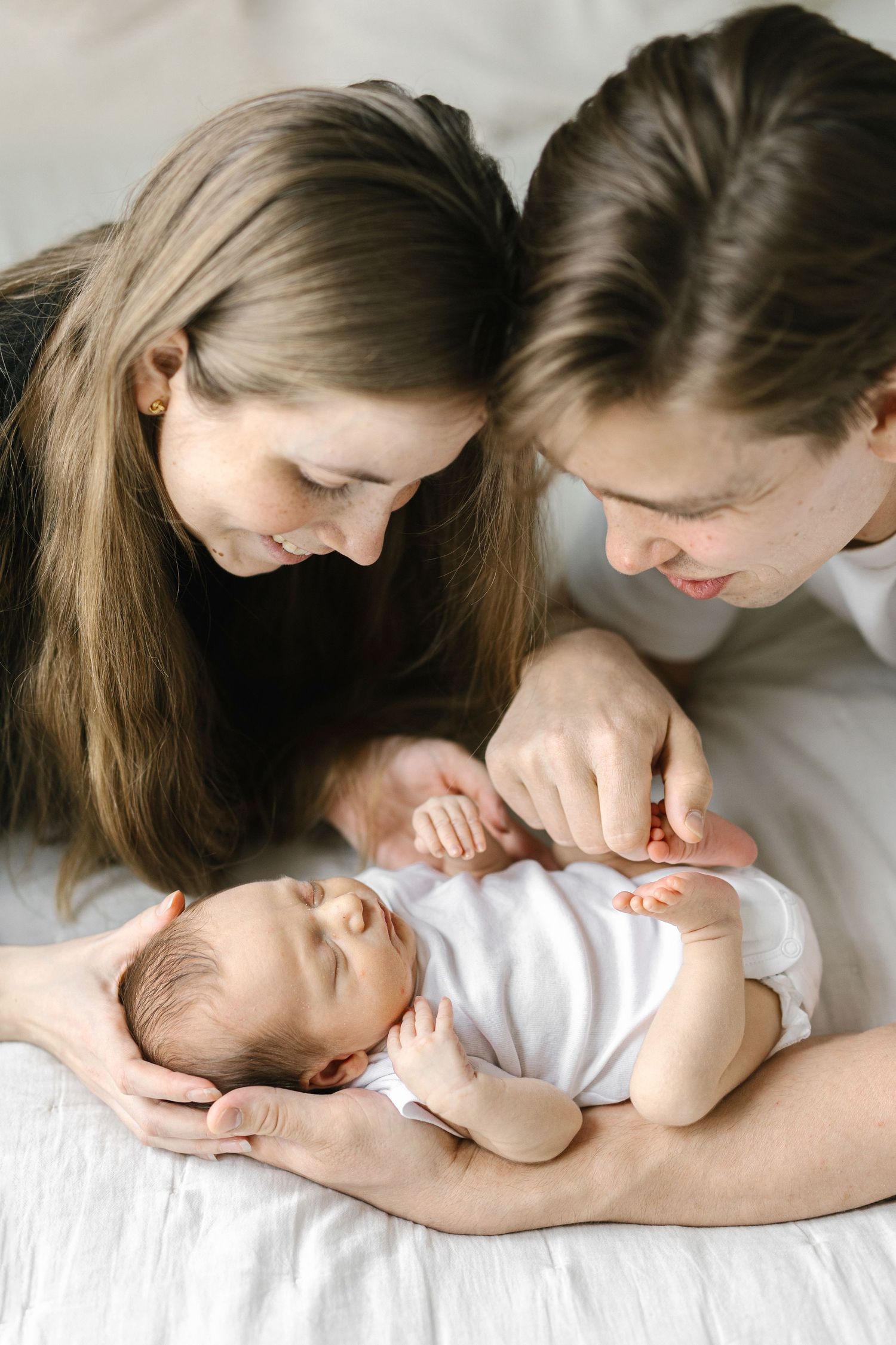 Two parents lovingly gaze at their sleeping newborn baby lying on a white bed.