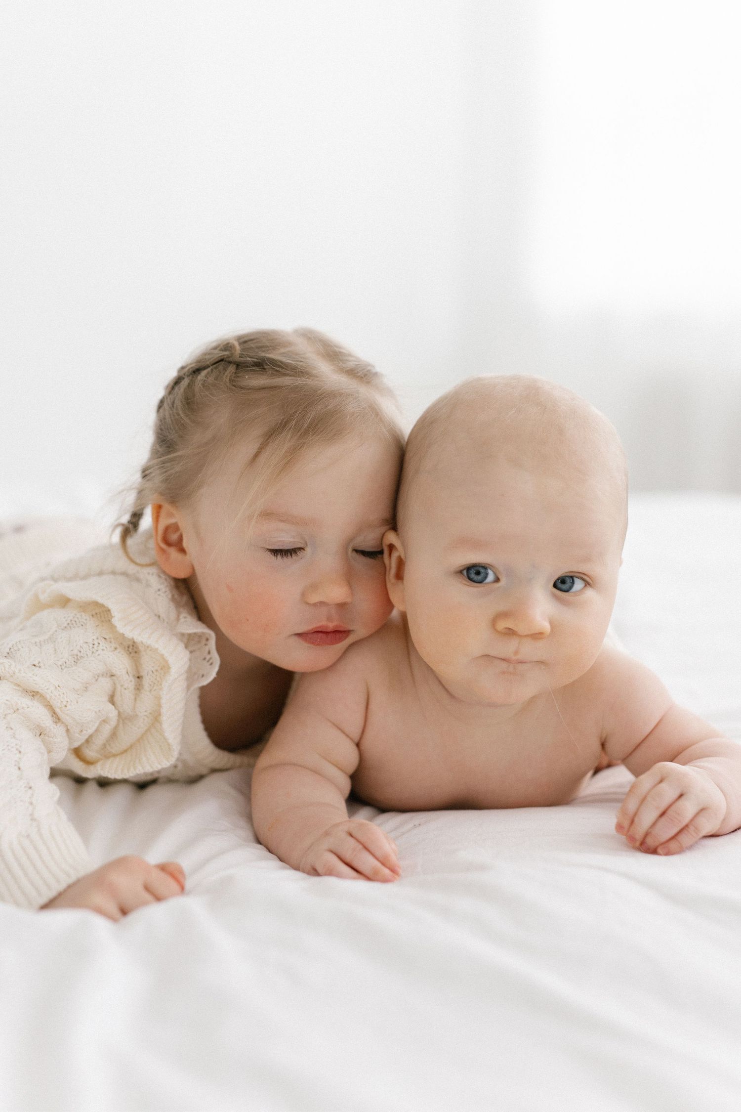 Toddler girl in cream sweater snuggles cheek-to-cheek with baby sibling on white bedding.