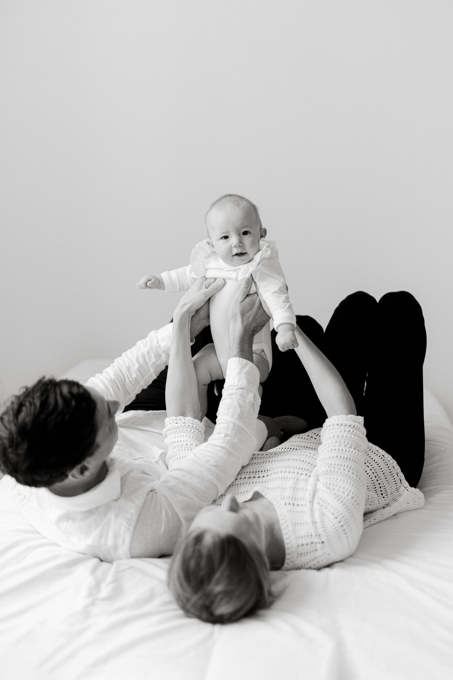 Black and white photo of parents lying on bed lifting their smiling baby in the air together.