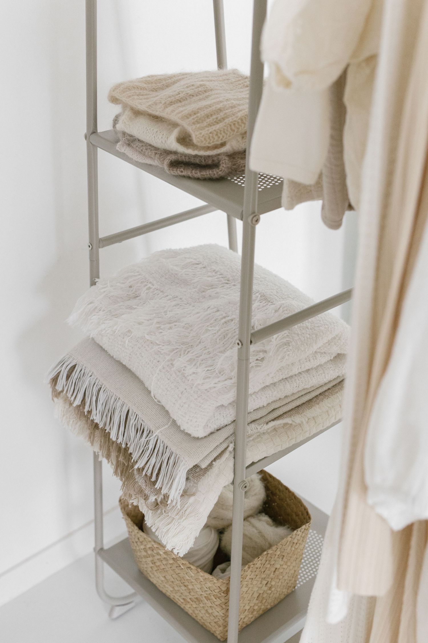 Metal ladder shelf with folded beige blankets, throws, and a wicker basket in a bright white minimalist room.