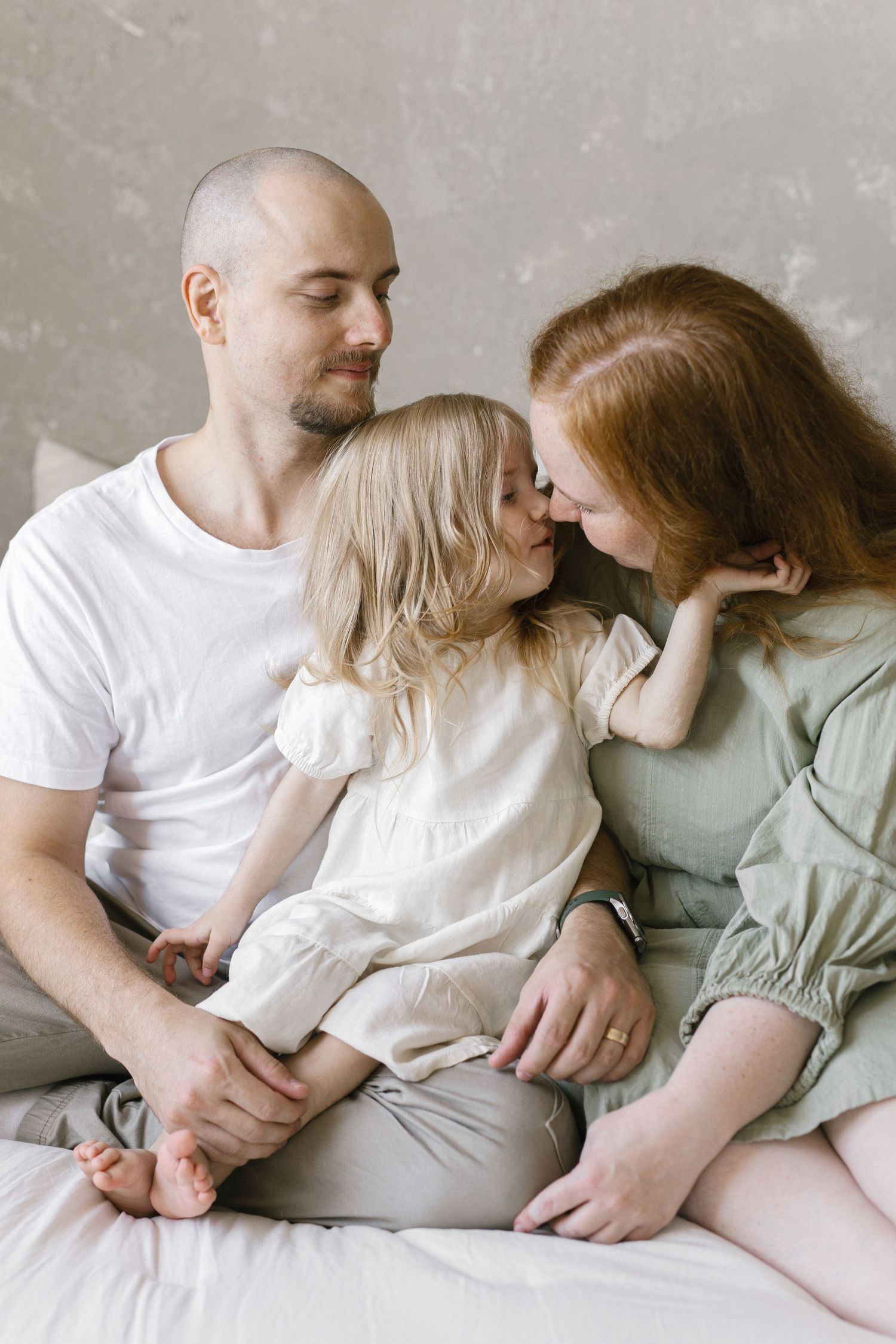 A loving family of three sits on a bed, with a blonde toddler kissing her redhead mother while father smiles warmly.