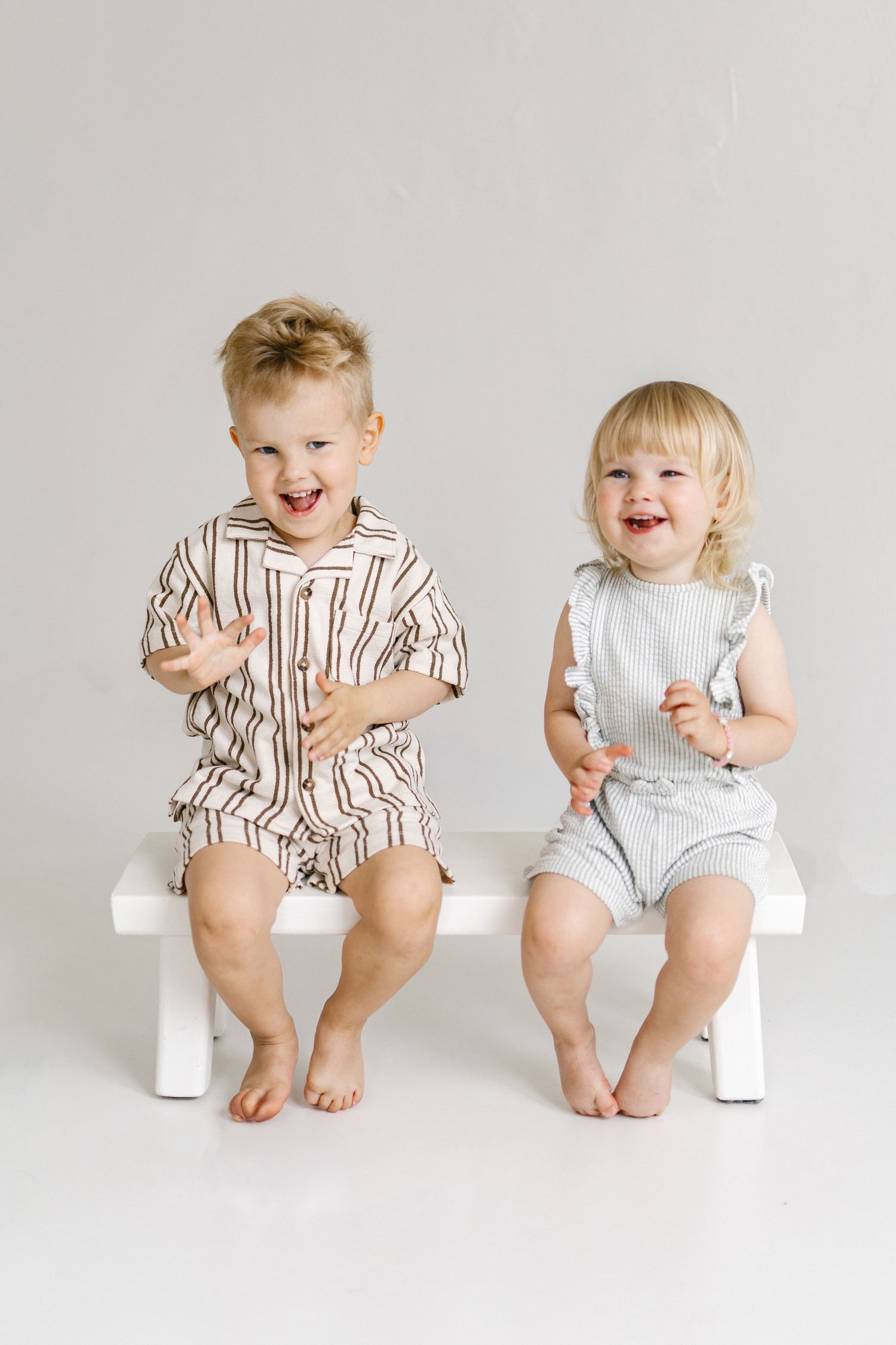 Two laughing toddlers sit on a white bench wearing matching striped and ribbed romper sets in neutral tones.