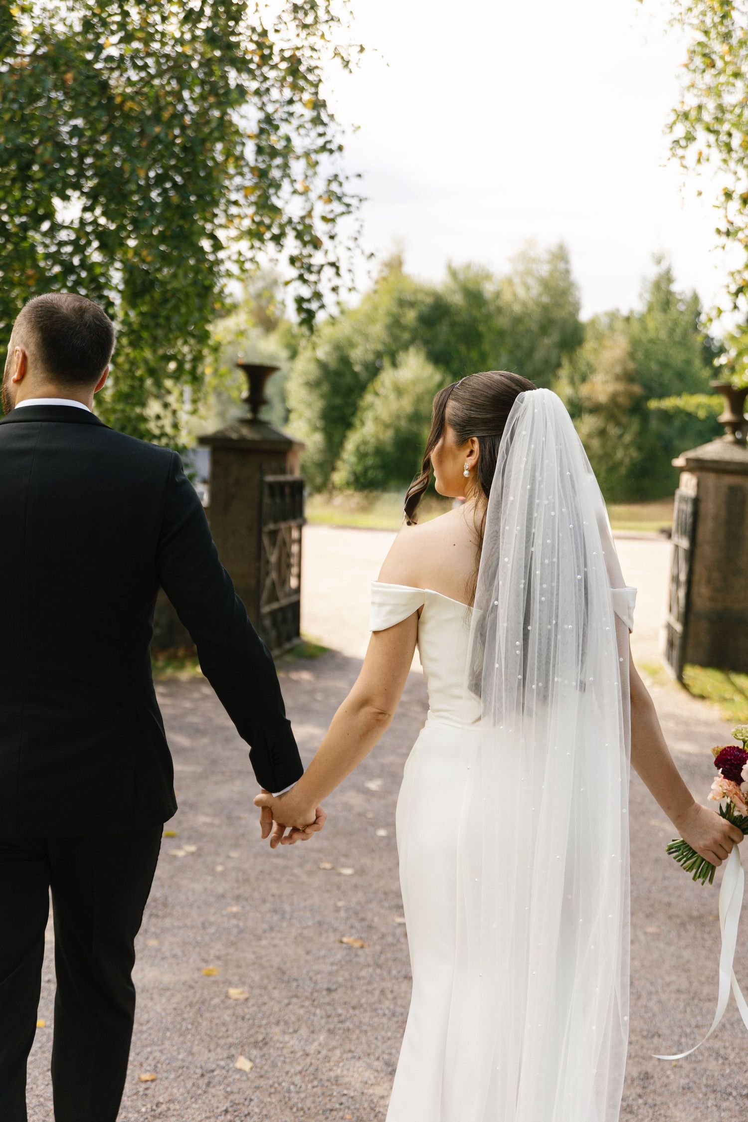 Bride and groom walking hand in hand through garden gates, bride wearing white off-shoulder gown with veil.