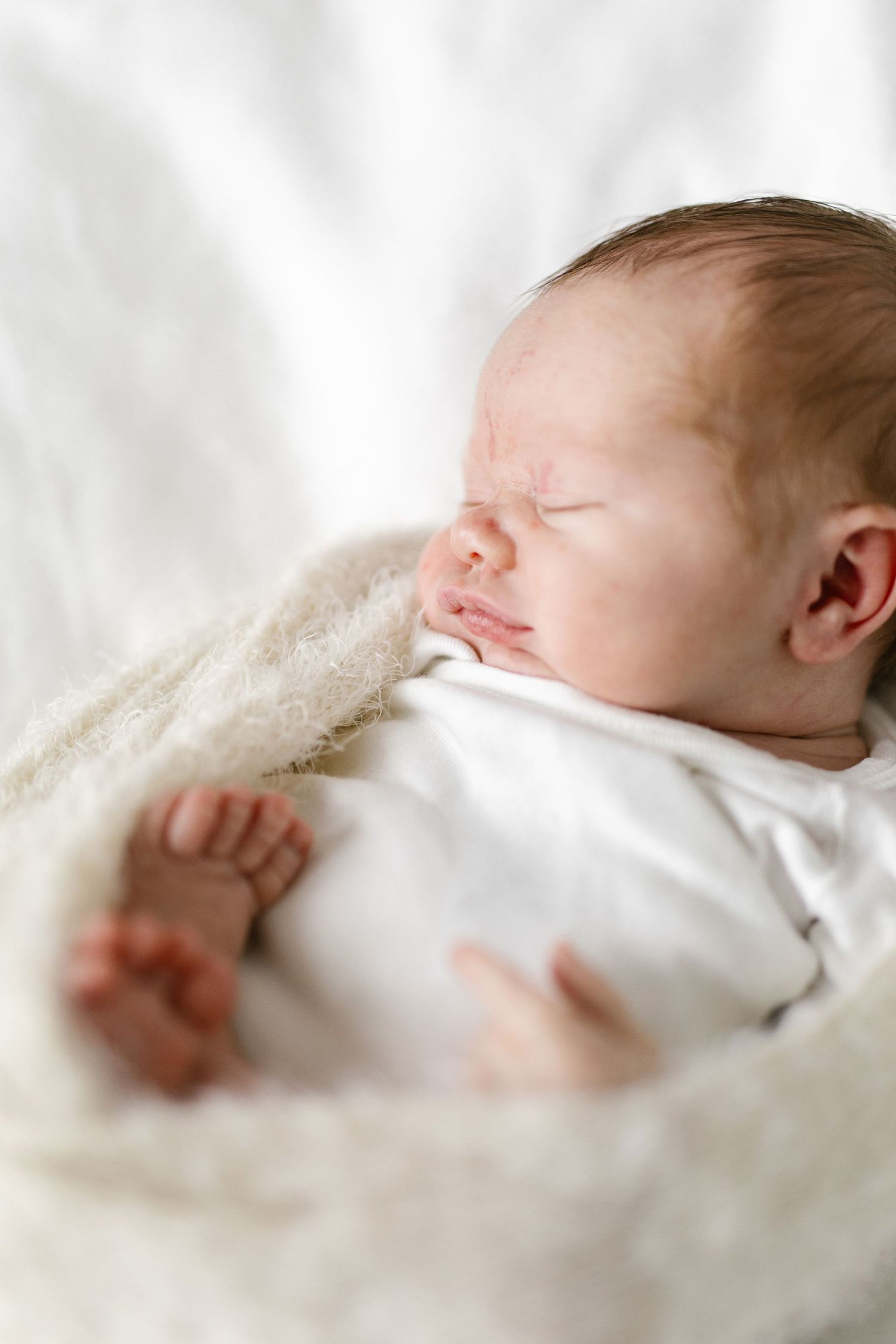 Peaceful newborn baby sleeping in soft white blankets with tiny feet visible.