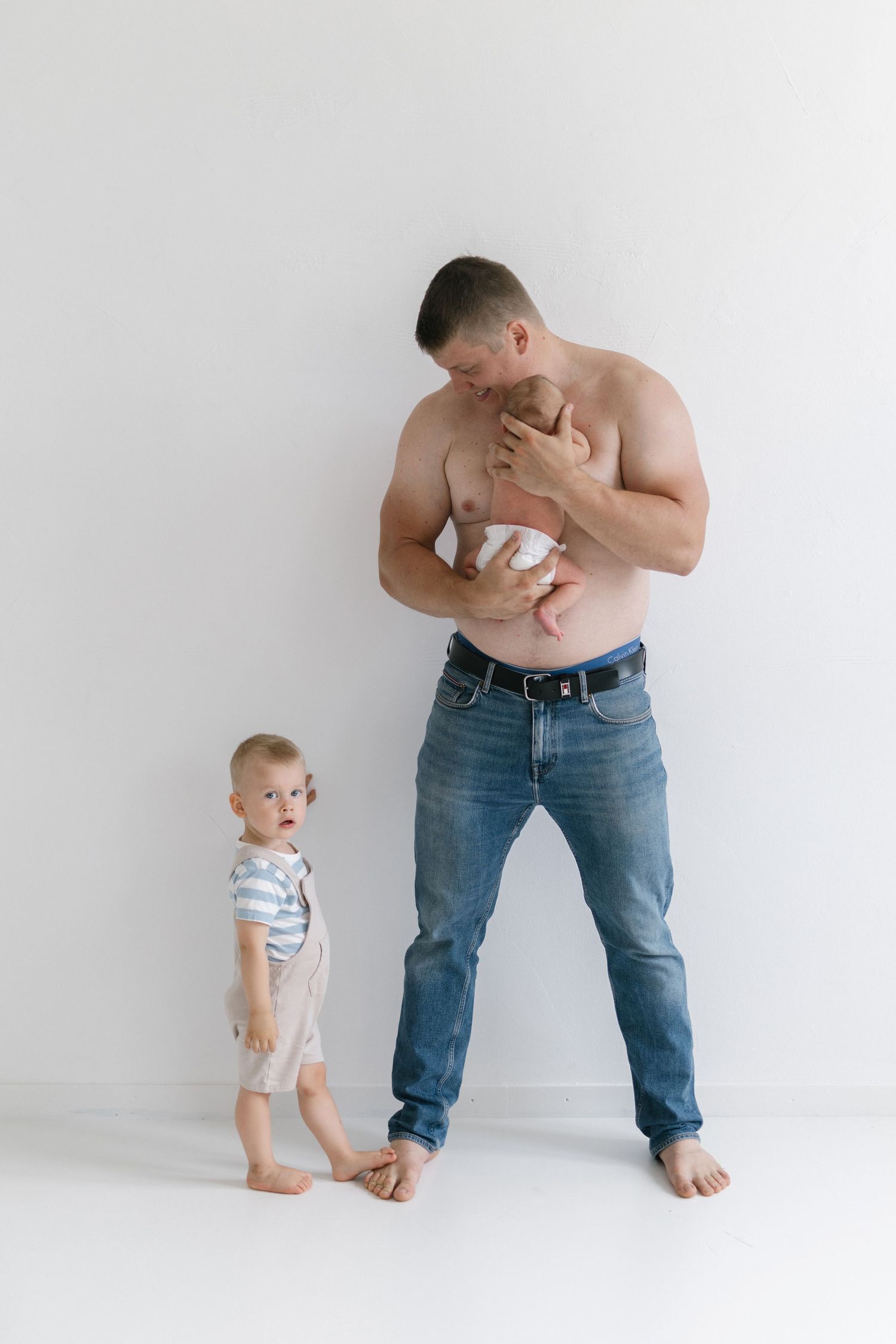 Parent holding newborn while toddler in striped shirt stands nearby against white background.