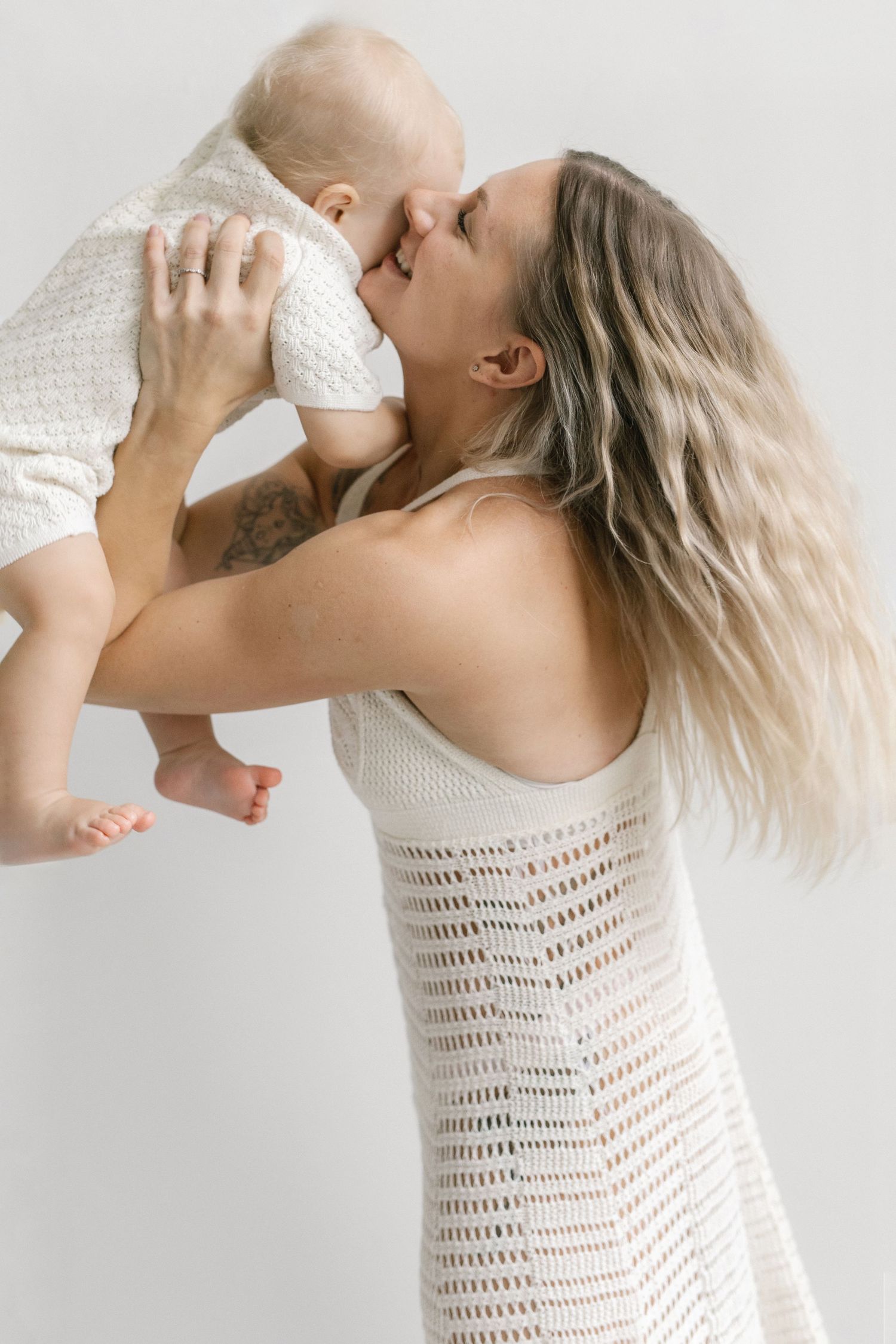 Smiling woman in a cream crochet dress lifts and kisses a baby in a white knit outfit against a soft neutral studio background.