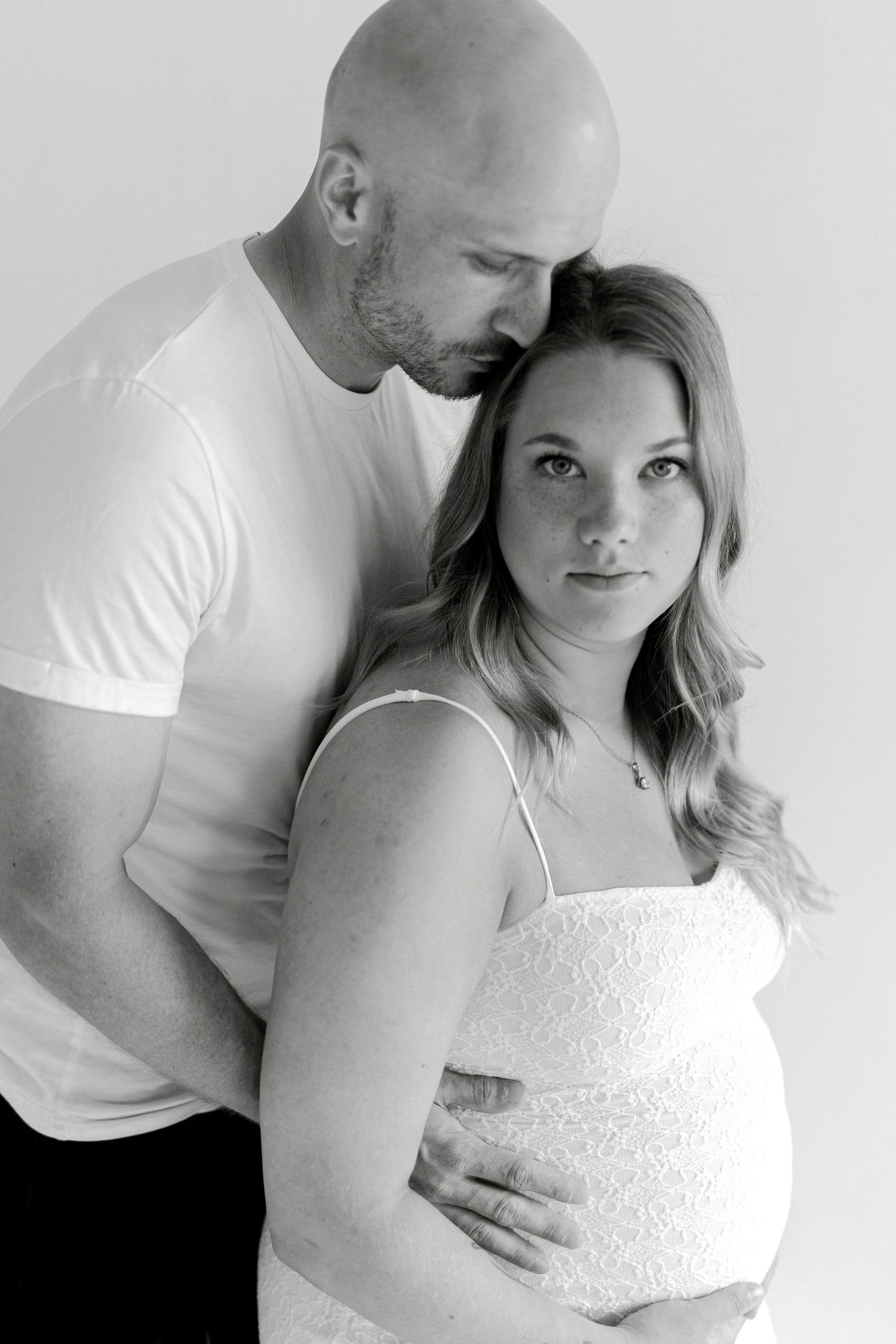 Black and white maternity portrait of a bald man kissing his pregnant partner's forehead as they cradle her baby bump.