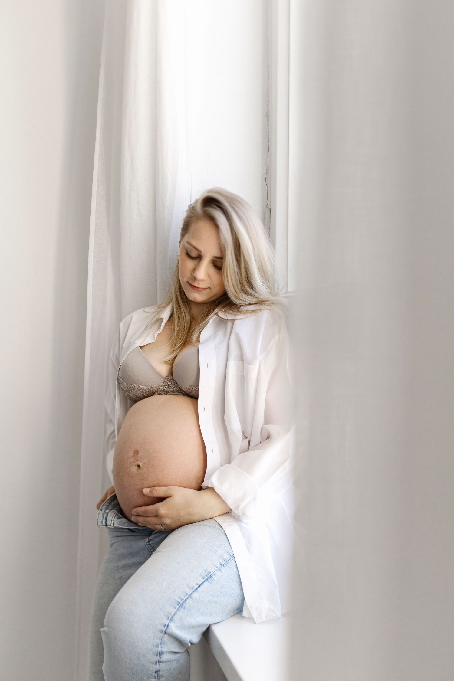 Pregnant woman in open white shirt and jeans cradling her bare baby bump by a bright window.