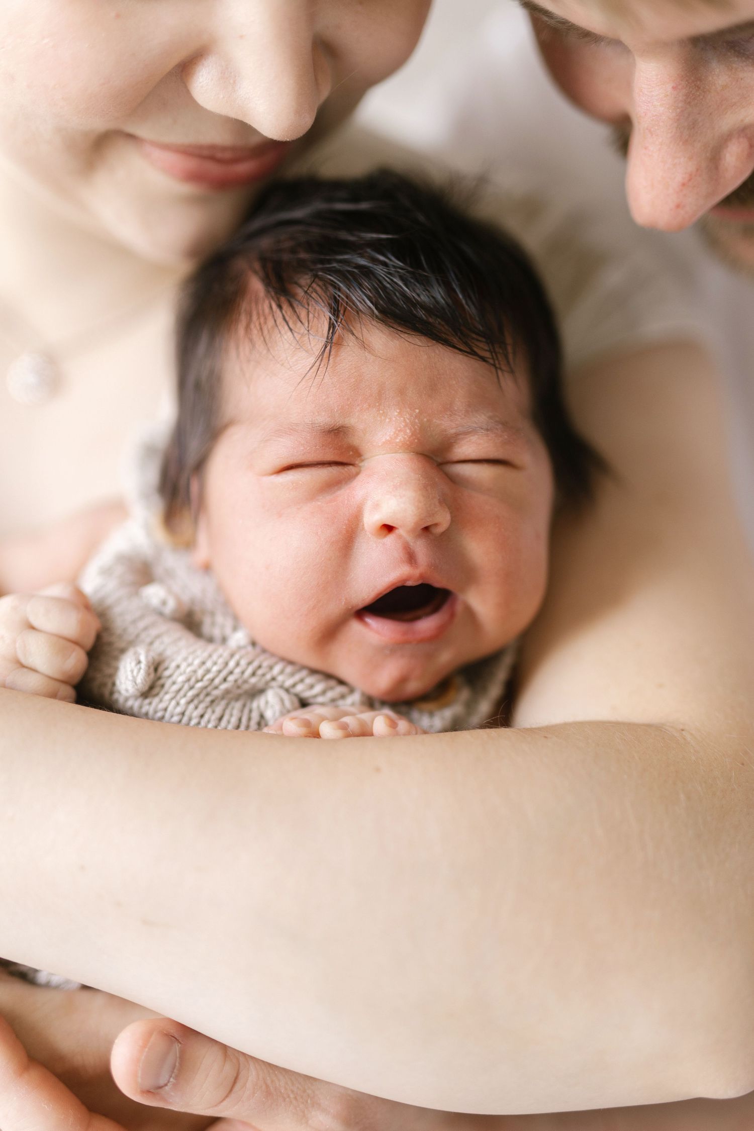 Two parents lovingly hold their newborn baby wrapped in a knit blanket, as the infant yawns contentedly.