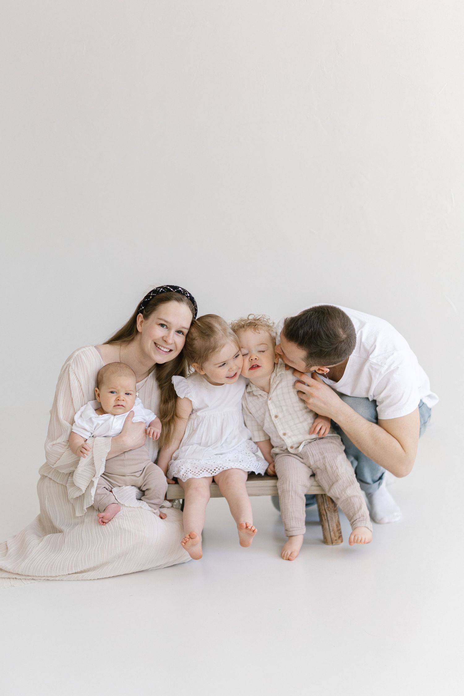 Happy family of five with mother holding newborn, father kissing toddler, and young children sitting together on white background.