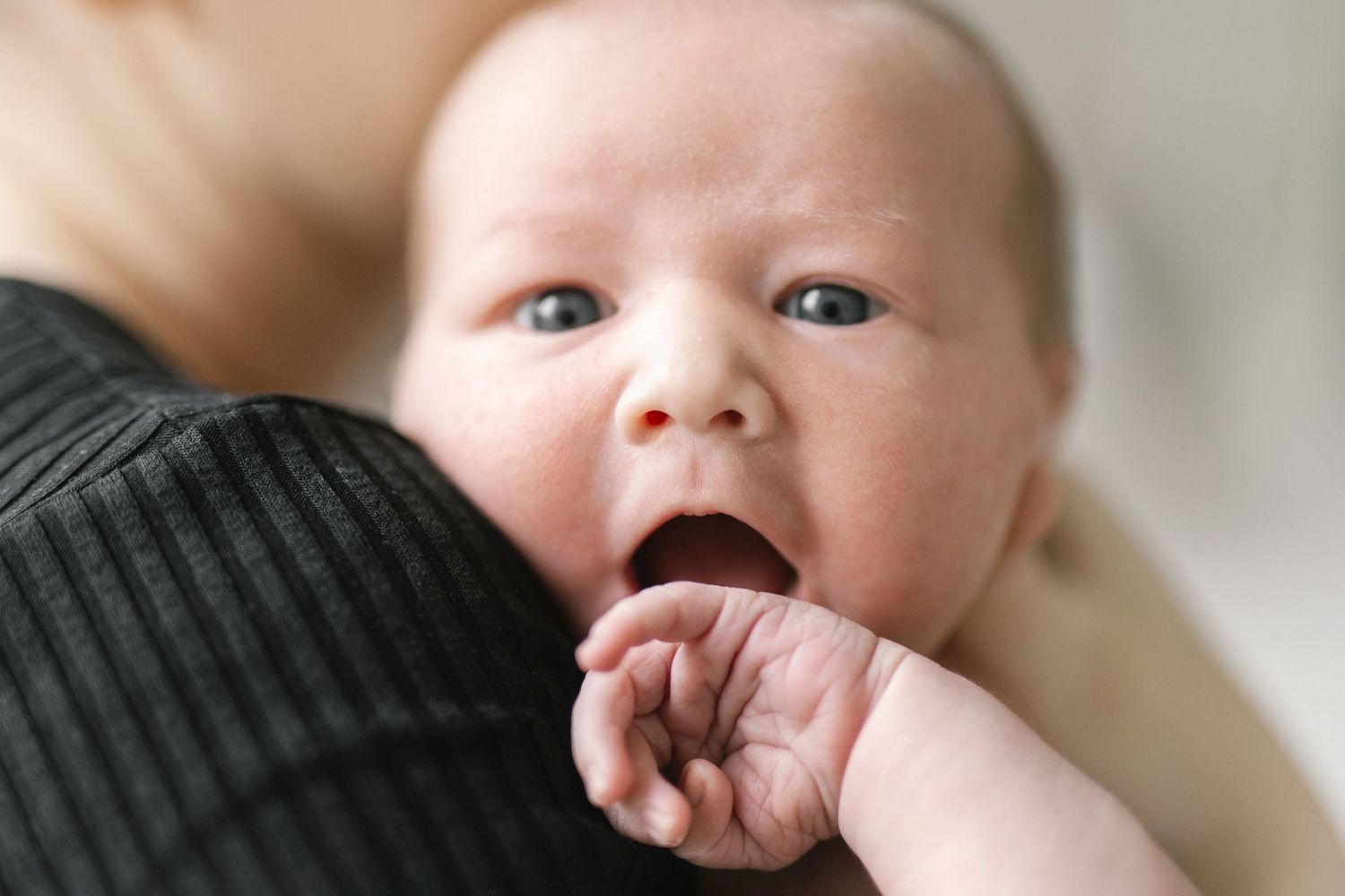 Close-up of a newborn baby with blue eyes, mouth open, chewing on tiny fingers while being held by a parent.