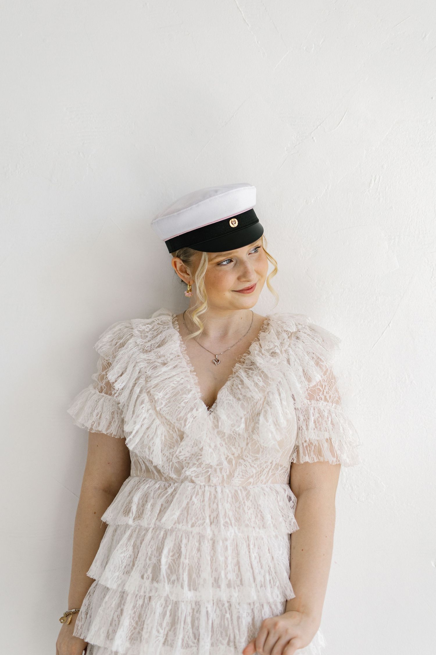 Blonde woman in white lace ruffle dress and white captain hat posing against a white wall.