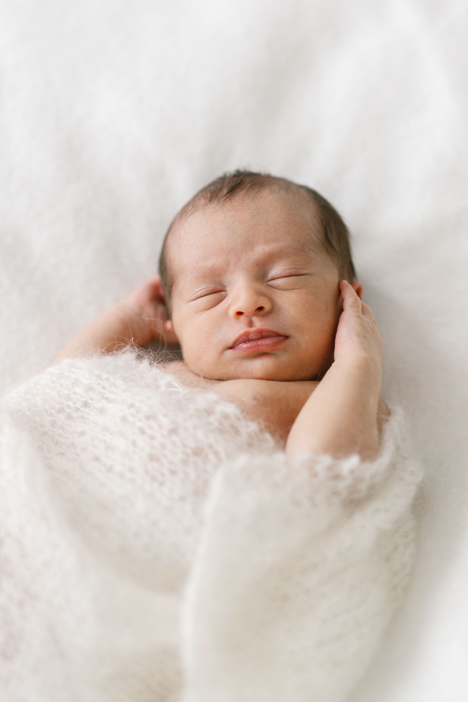 Peaceful newborn baby sleeping wrapped in a soft white knit blanket with tiny hands tucked under chin.