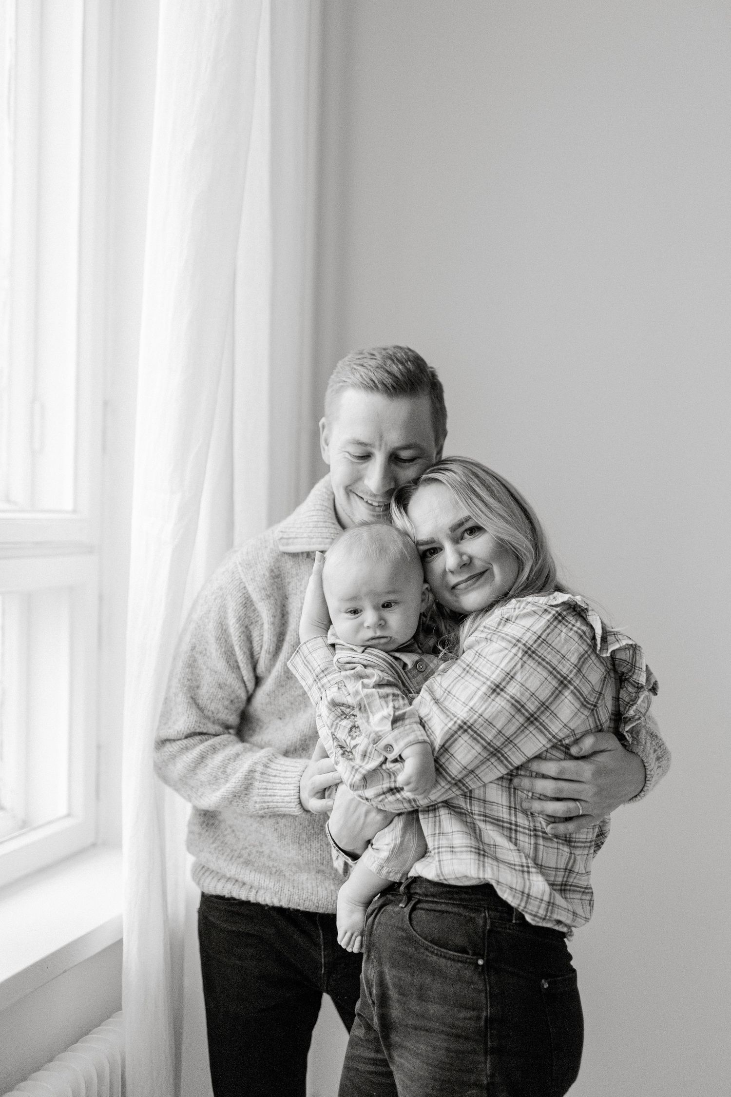 Black and white photo of a happy young family with parents smiling and holding their newborn baby by a bright window.