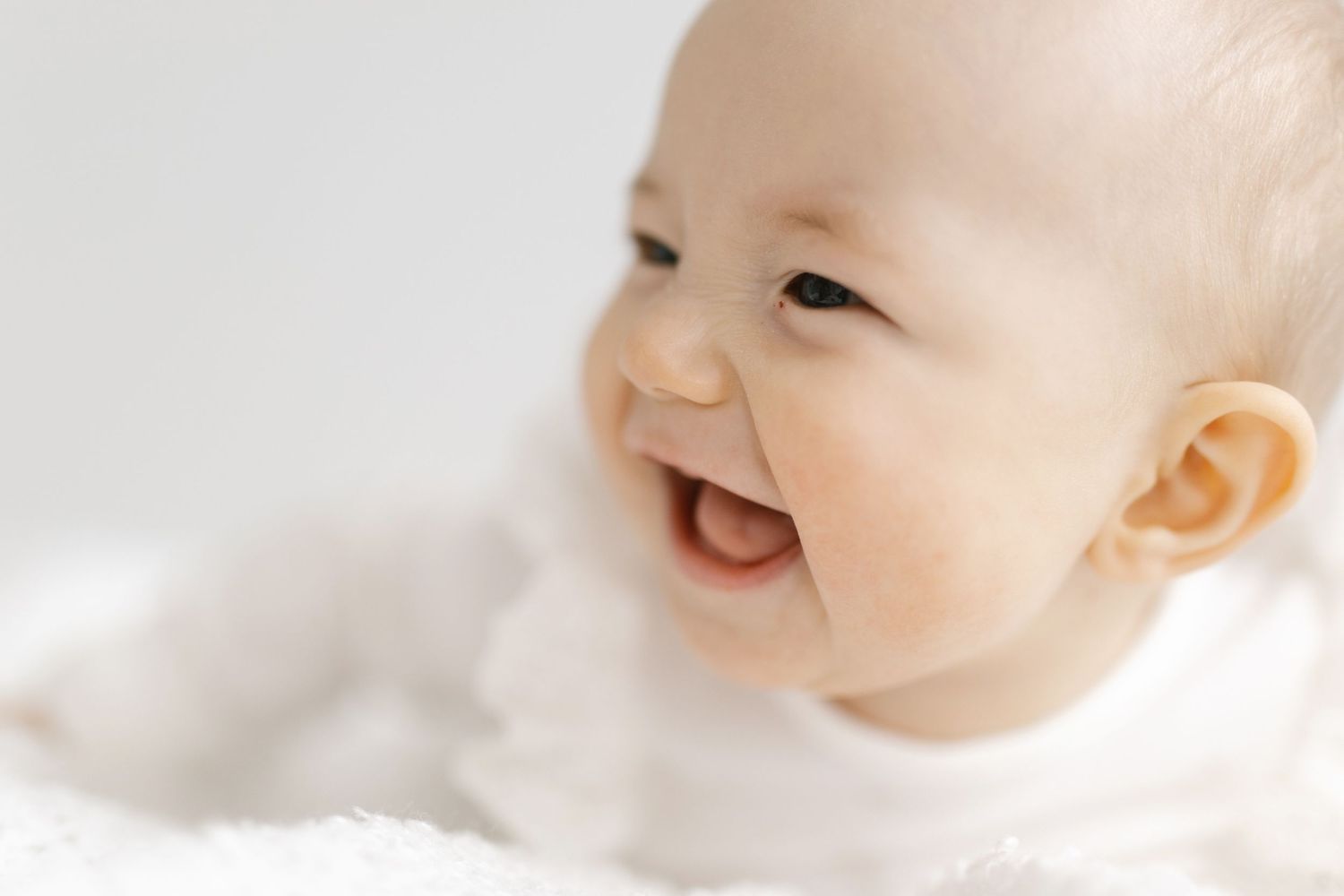 Adorable smiling baby lying on a soft white blanket, looking to the side with a joyful expression.