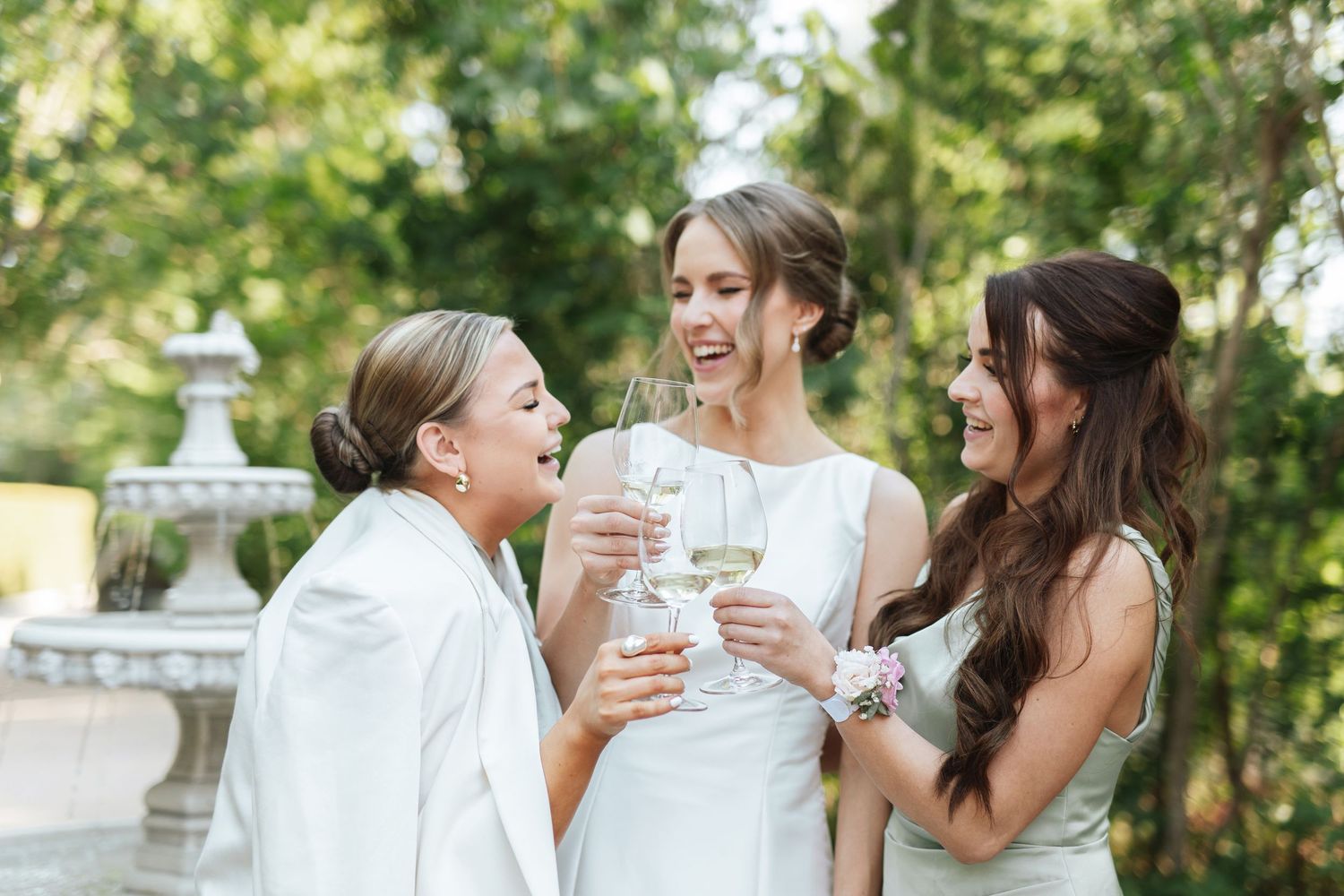 Three women in white dresses toasting with champagne glasses outdoors at a wedding celebration.