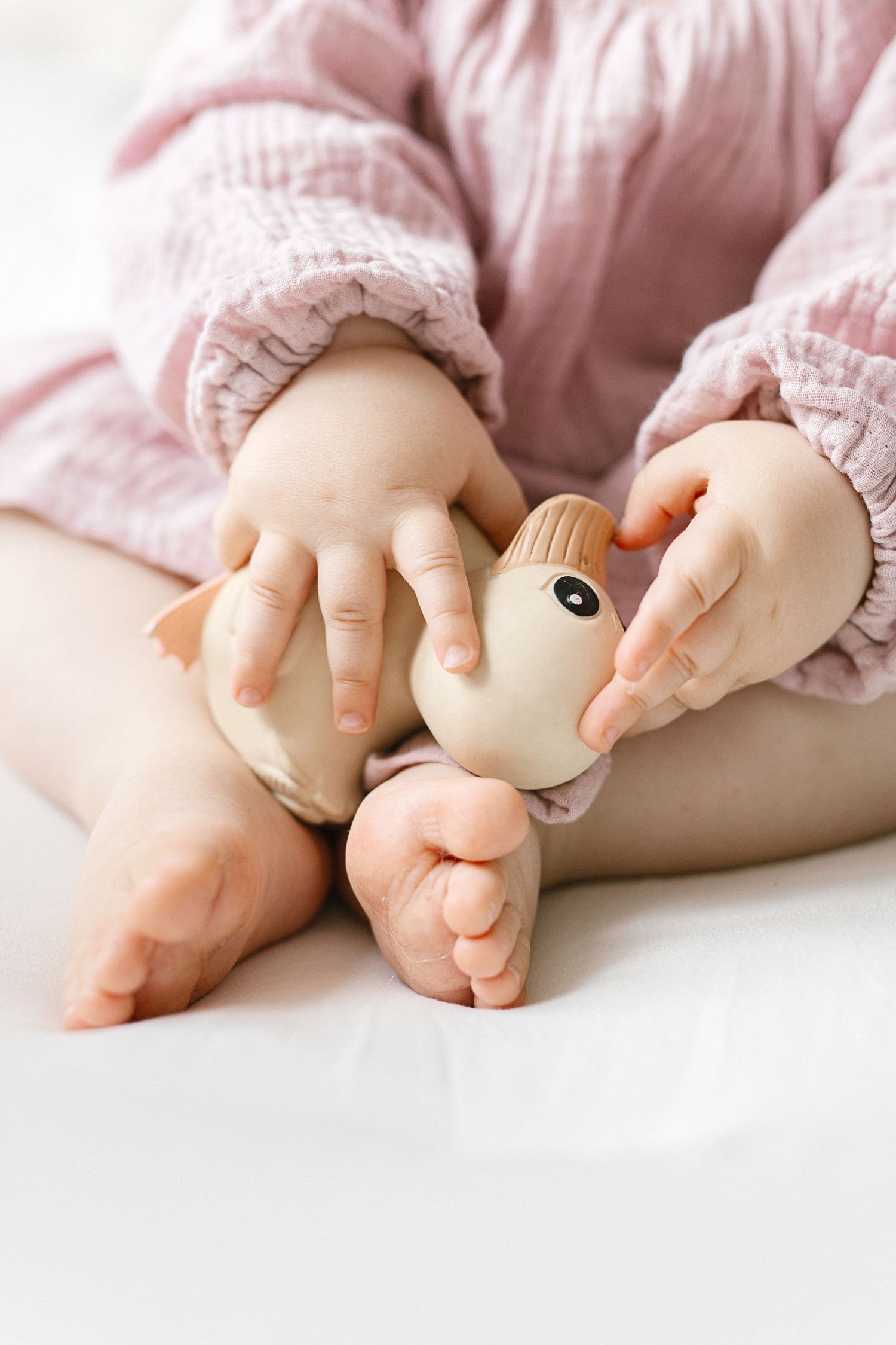 Baby in pink outfit holding a rubber duck toy while sitting, with tiny bare feet visible in foreground.