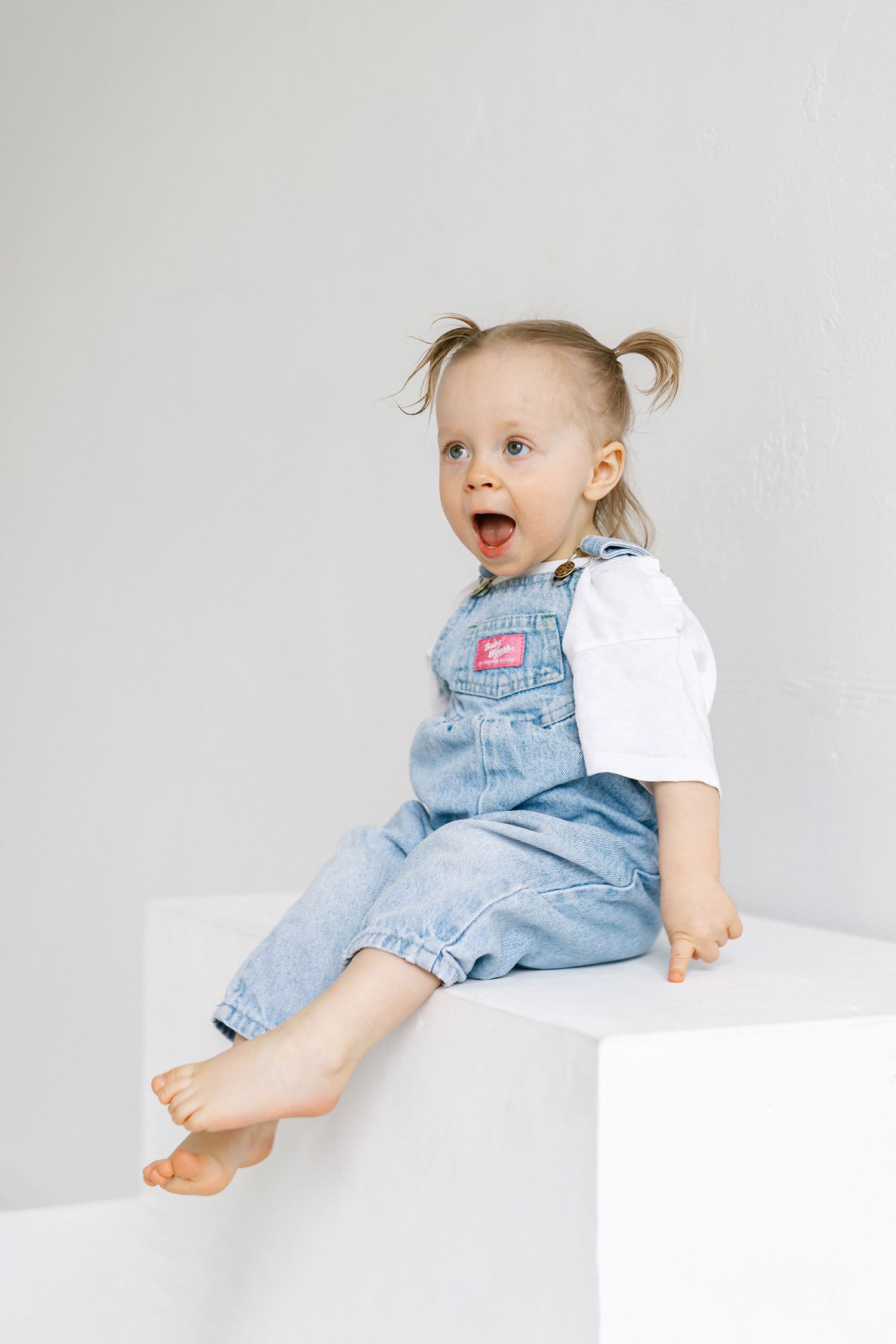Cute toddler girl with pigtails sitting on white cube, wearing denim overalls and white shirt, mouth open in surprise.