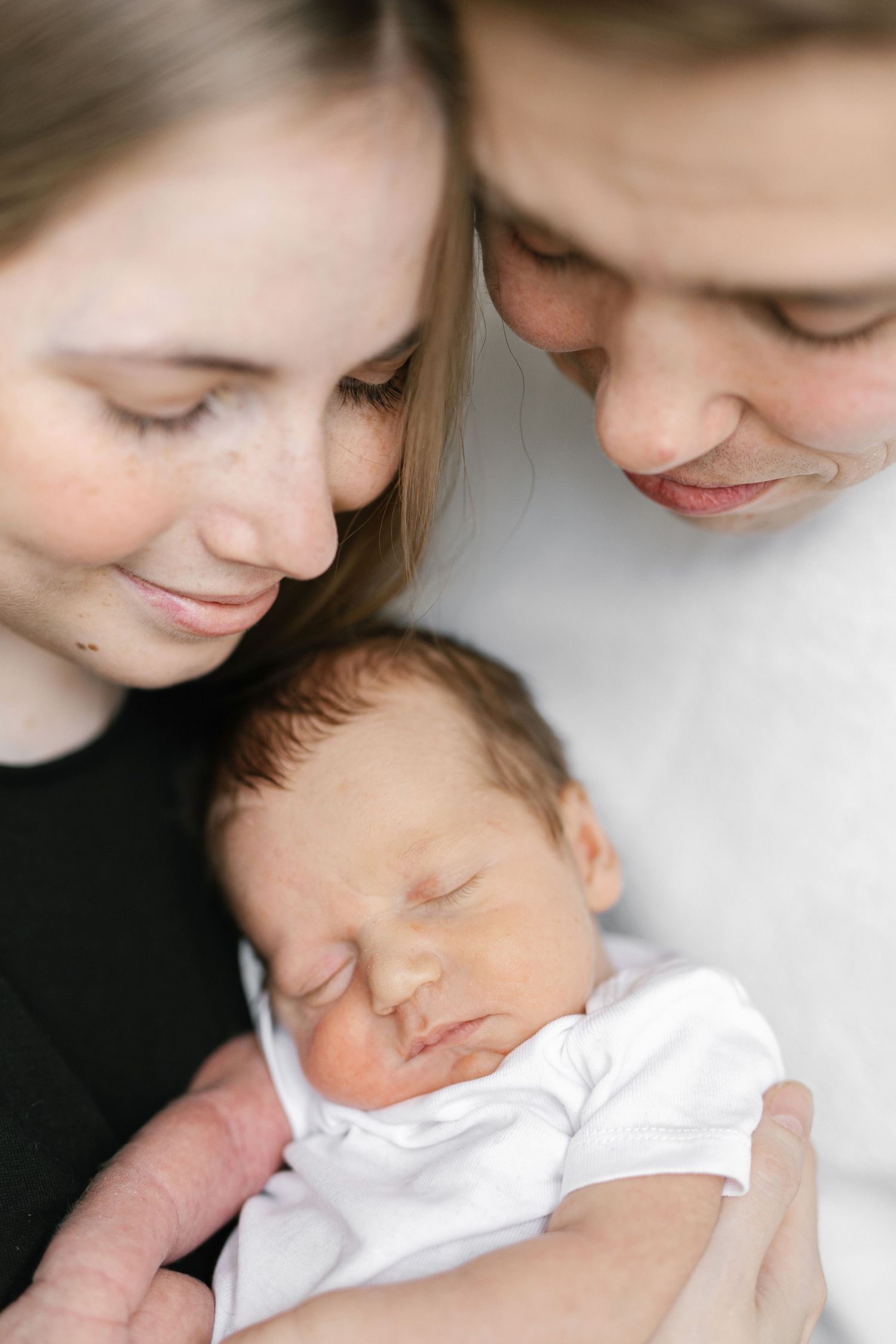 Two parents lovingly gaze at their sleeping newborn baby dressed in white, in a tender family portrait.