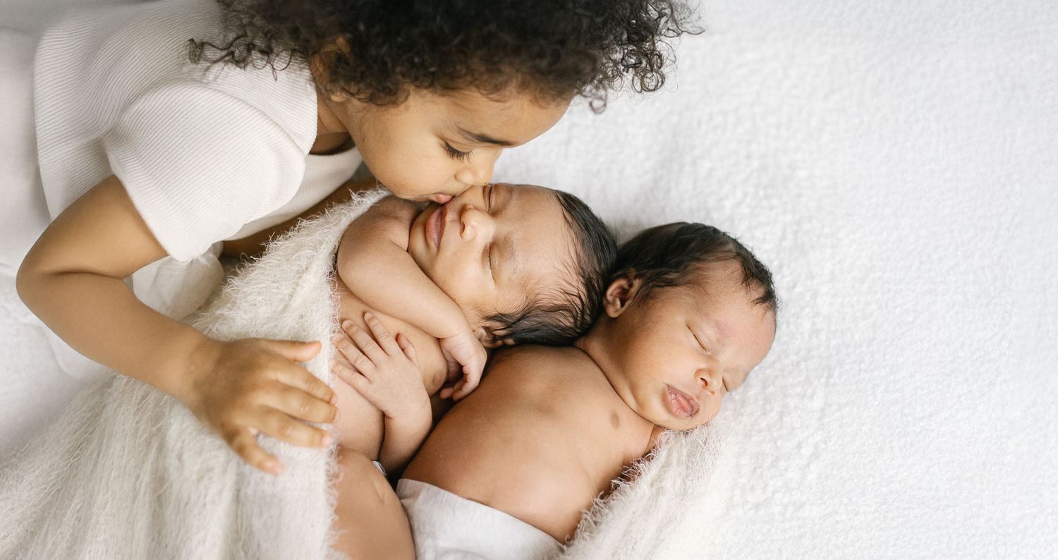 A young child lovingly kisses newborn twins wrapped in white blankets on a soft white bed.