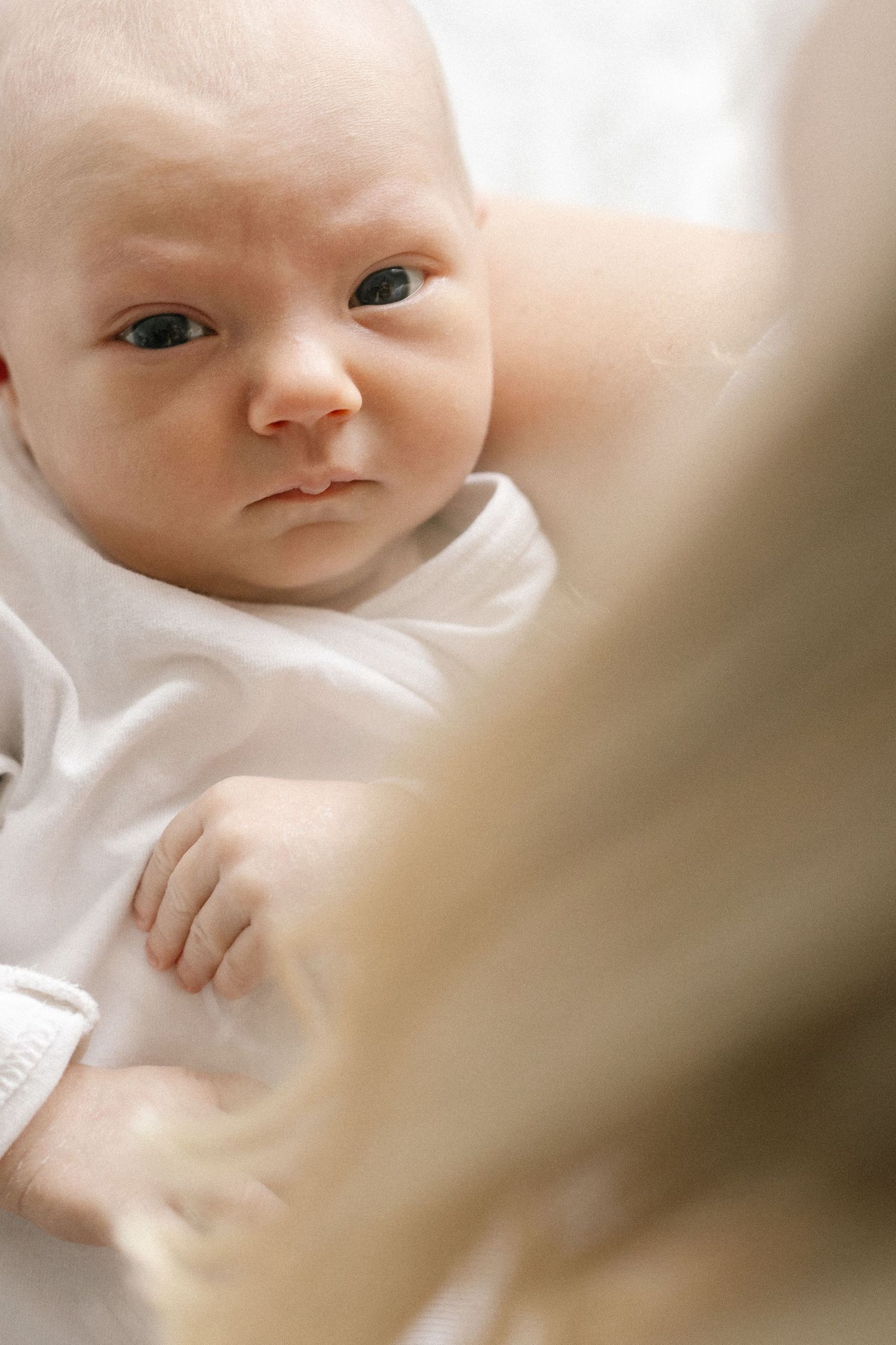 A newborn baby in a white outfit gazes upward with wide eyes while being cradled in an adult's arms.