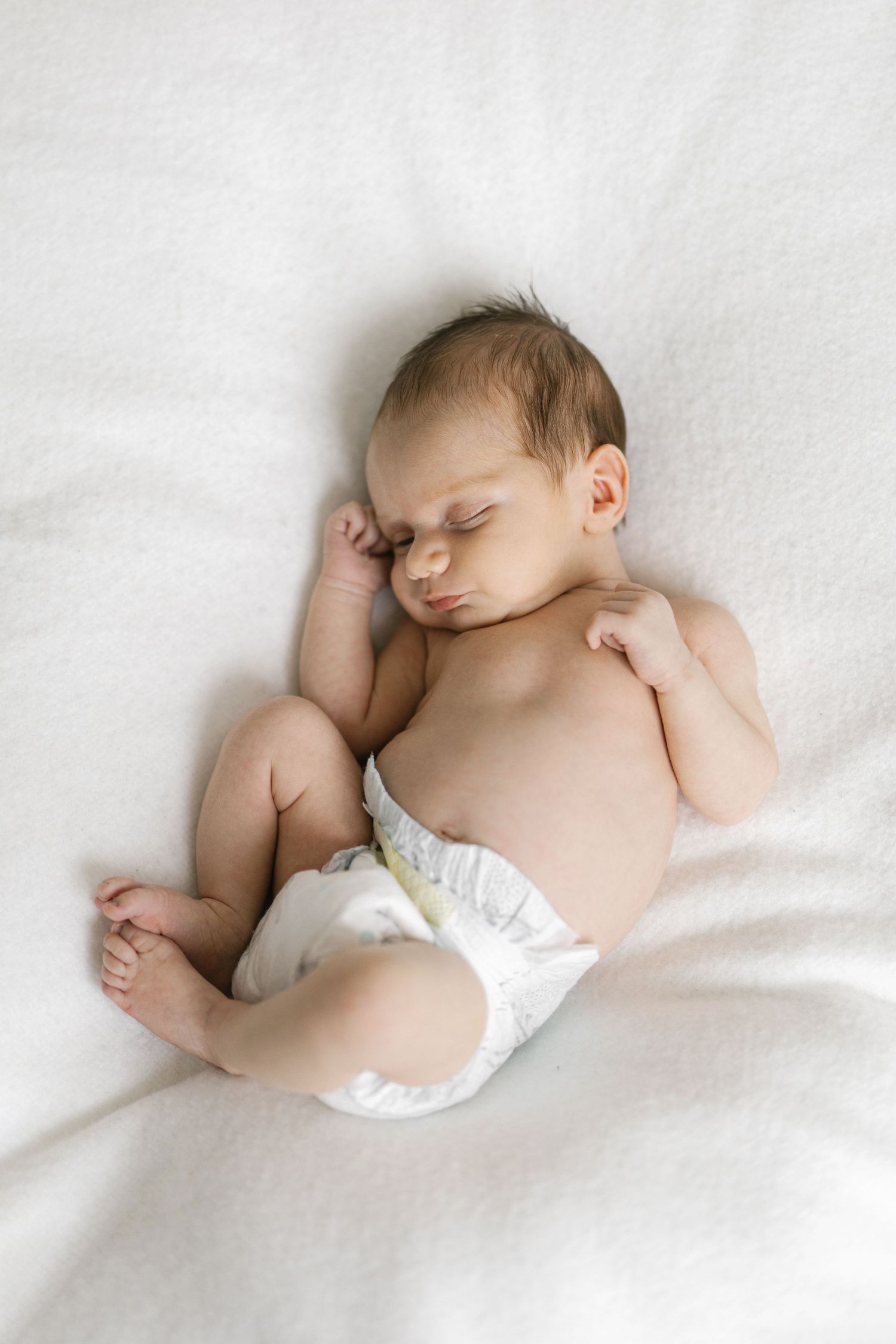 Peaceful newborn baby sleeping on white blanket wearing diaper, curled in natural position with tiny fists and feet visible.