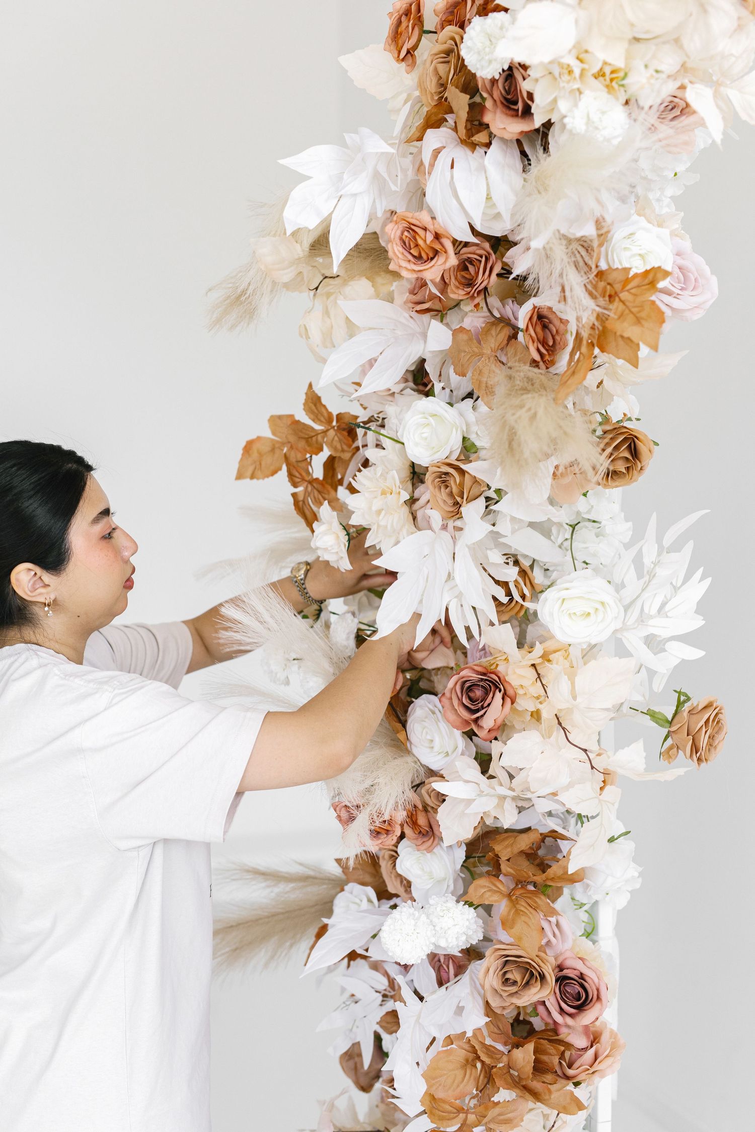 Woman in white arranging a lush floral arch with roses, white blooms, and golden leaves for a wedding.