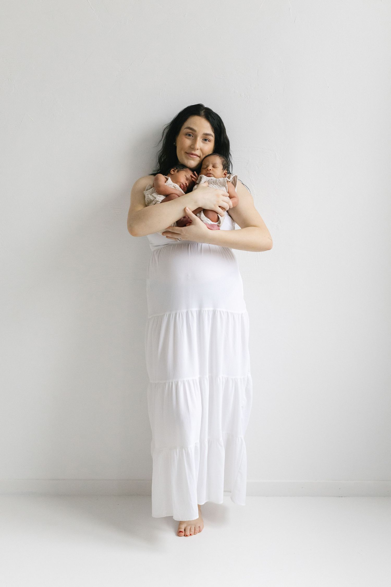 A woman in a white maxi dress holds two newborn babies against a white background.