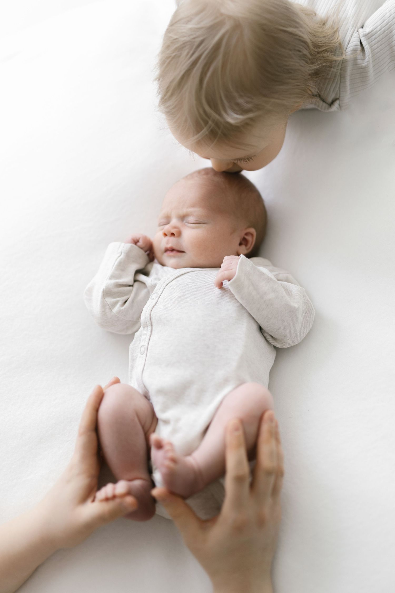 Toddler gently kissing sleeping newborn sibling in white onesie.