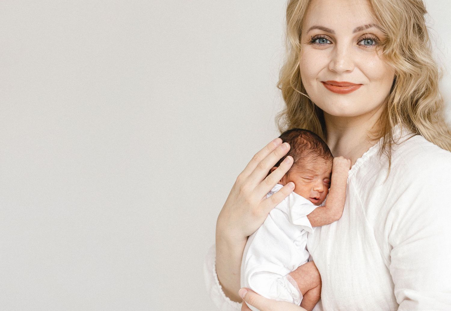 Smiling blonde woman in white top tenderly holding a sleeping newborn baby against a light gray background.