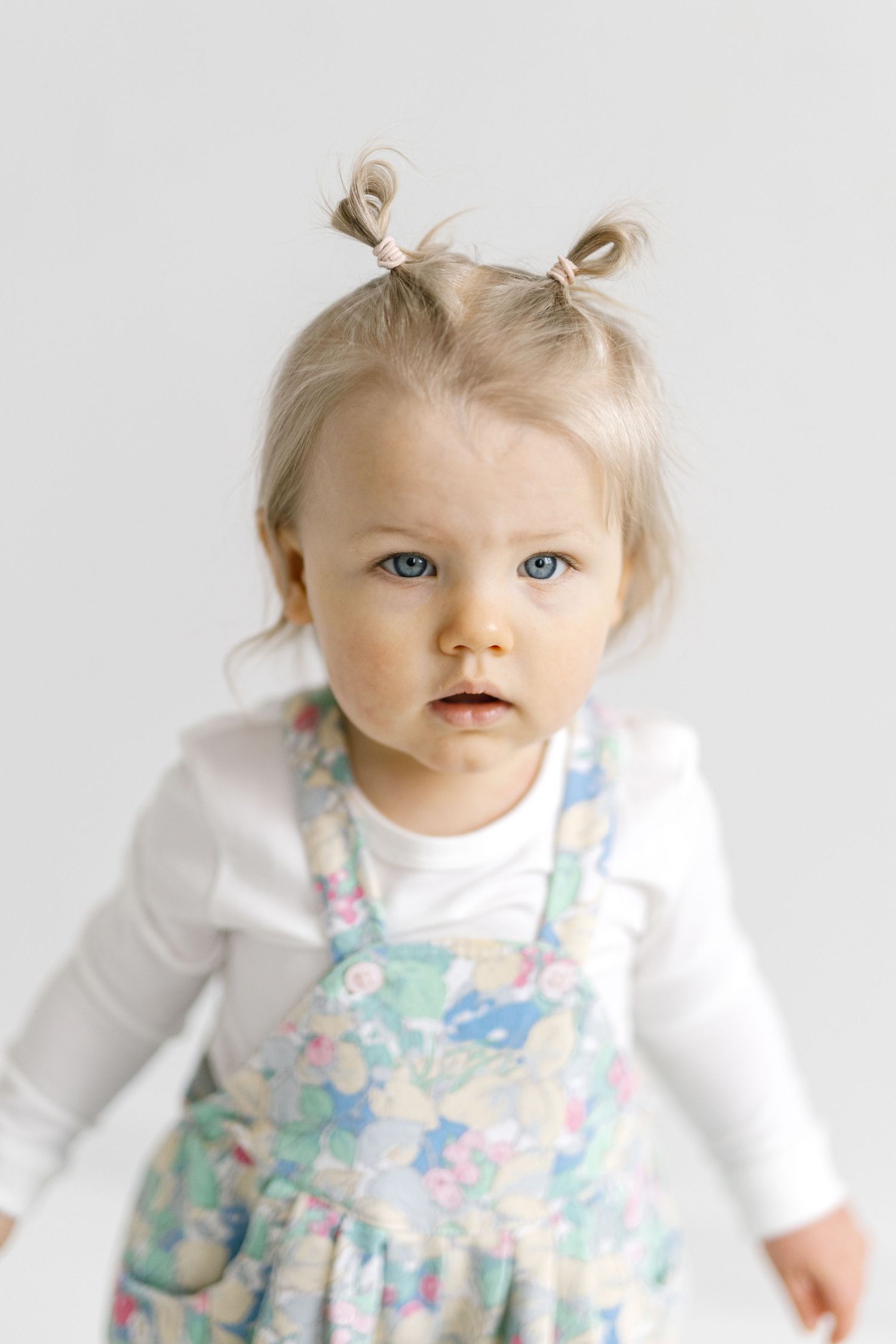 Adorable blonde toddler girl with pigtails and blue eyes wearing a floral dress over a white shirt.
