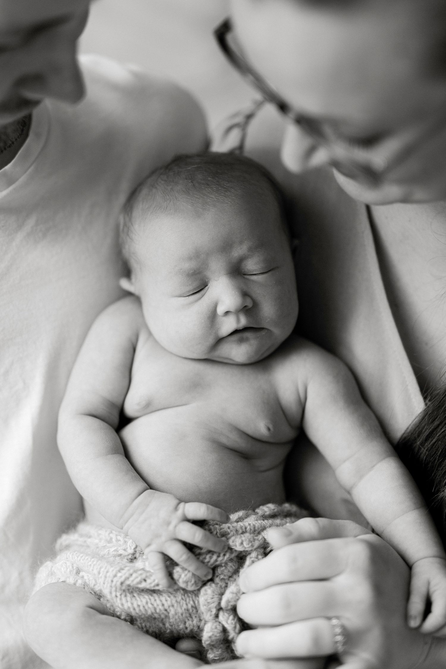 Black and white photo of a sleeping newborn baby cradled in a parent's arms, wrapped in a knit blanket.