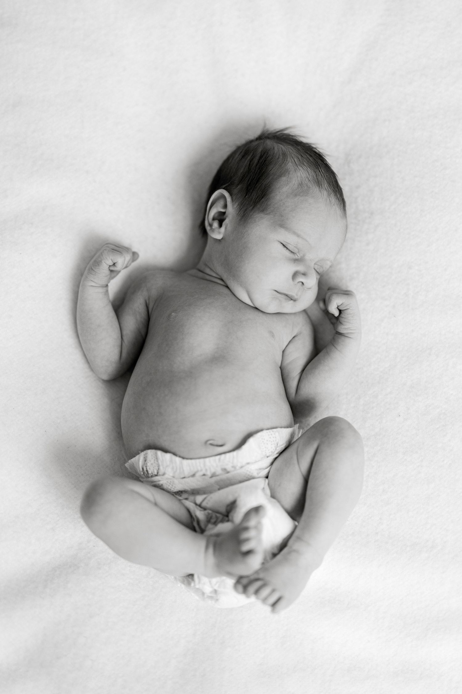 Black and white photo of a sleeping newborn baby in a diaper lying on a soft white blanket.
