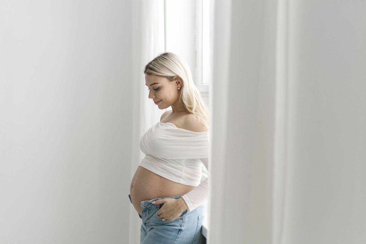 Pregnant woman in white off-shoulder top and jeans gently holding her baby bump near a bright window.