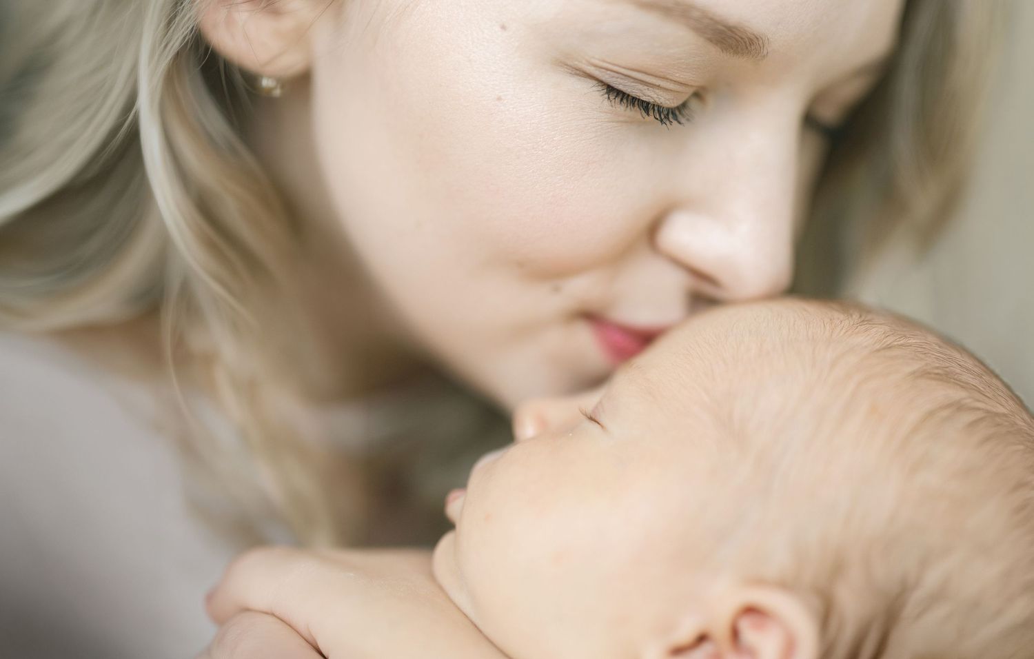 A blonde mother with closed eyes gently kisses the forehead of her sleeping newborn baby in a soft, tender moment.