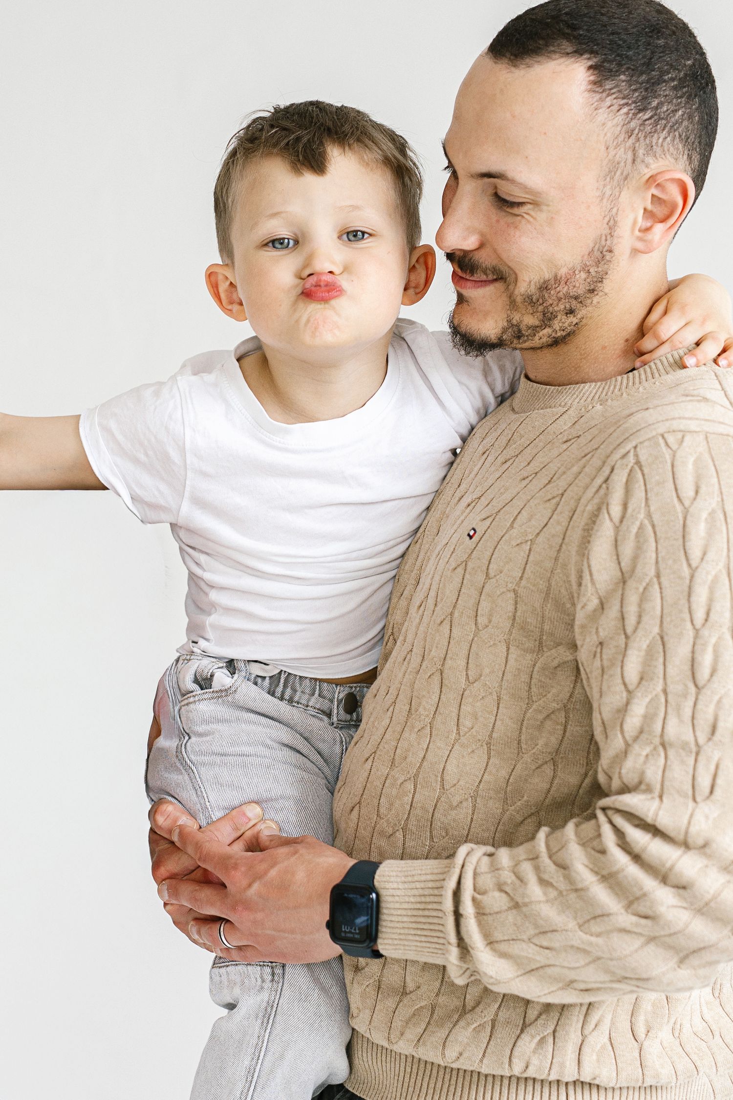 A bearded father holds his young son who makes a funny face, both wearing casual clothes against a white background.
