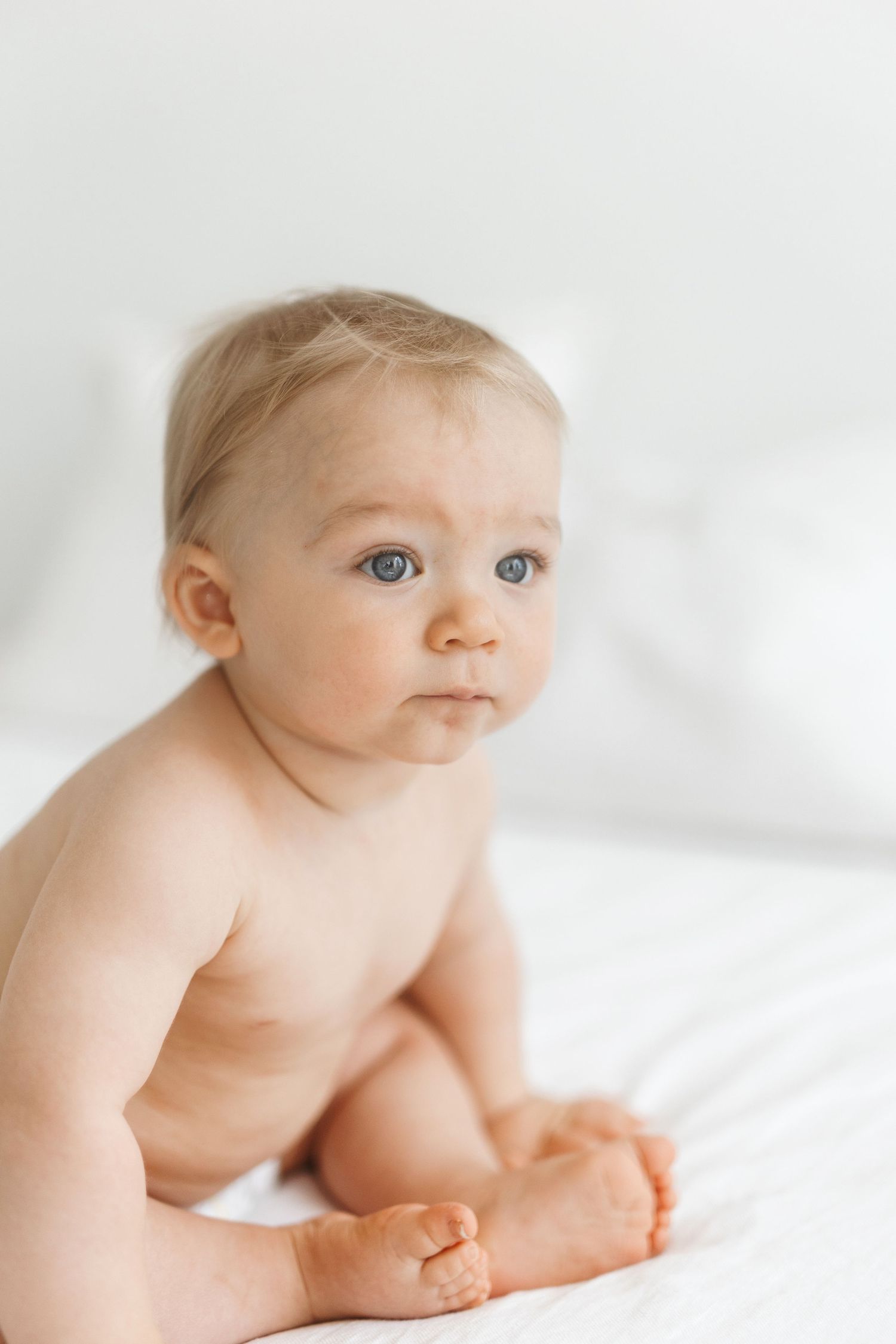Cute baby with blue eyes sitting on white bed, looking curiously to the side in soft natural light.